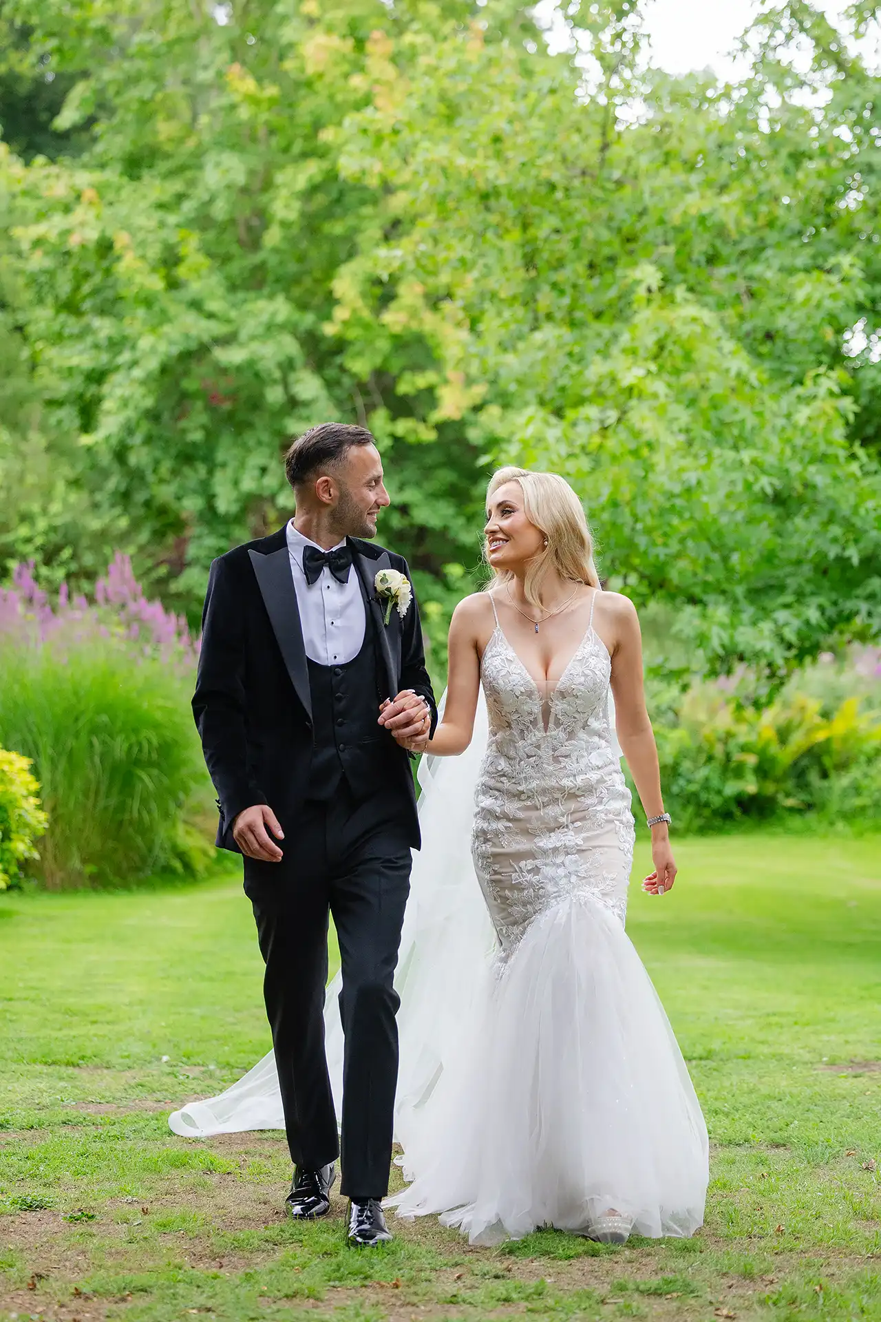 bride and groom walking Bride in a white lace wedding dress and groom in a black tuxedo walk hand in hand through a lush green garden.