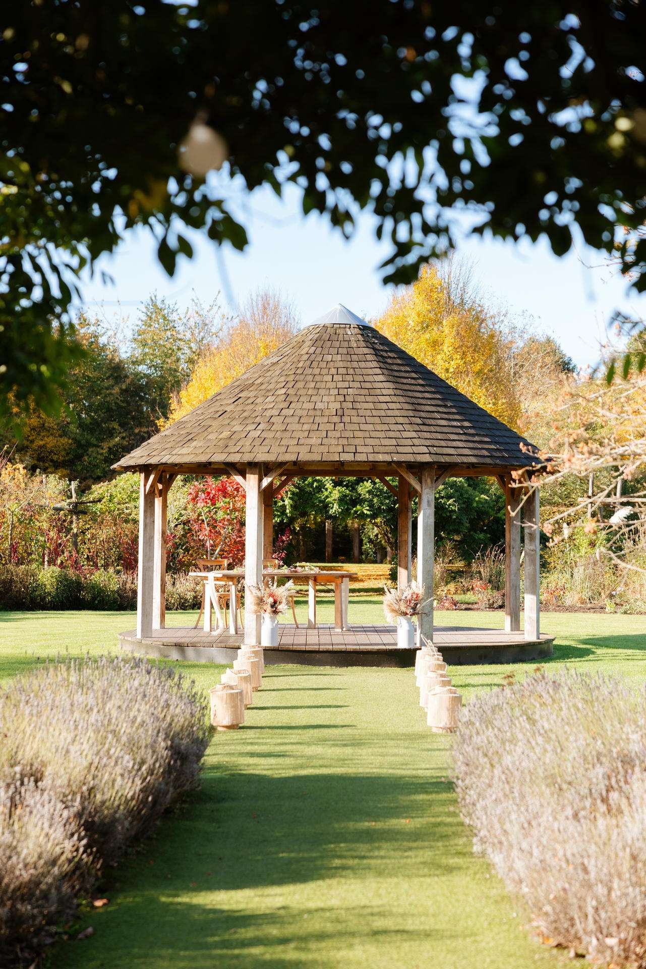 A wooden gazebo with a conical shingled roof sits in a well-kept garden, with a grassy path lined by lavender leading up to it and autumn trees in the background.