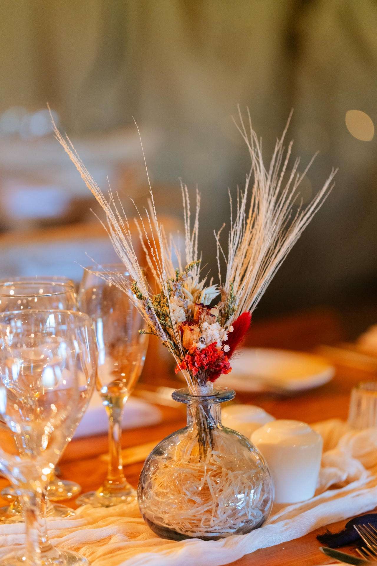 Round glass vase with a dried floral arrangement sits on a candlelit dinner table beside wine glasses.