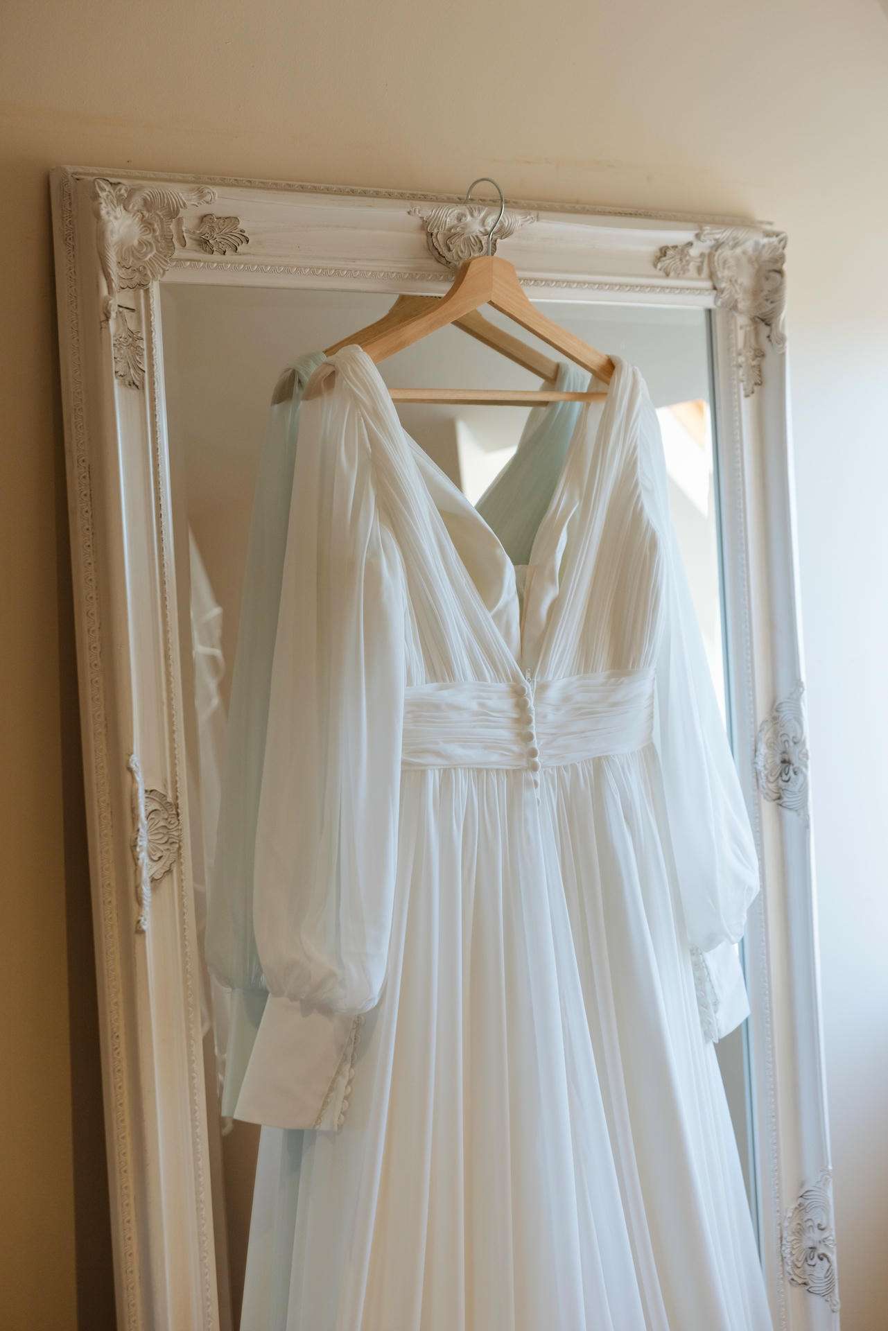 A white wedding dress with sheer puff sleeves hangs on a wooden hanger in front of an ornate white-framed mirror.