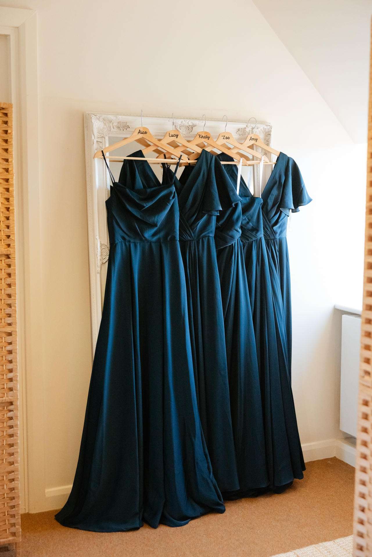 Five teal bridesmaid dresses on wooden hangers hung on a white ornate frame against a pale wall.