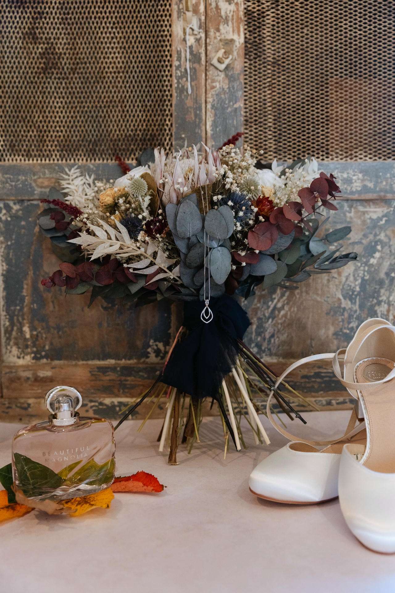 A dried bouquet of eucalyptus and muted beige, cream, and burgundy flowers sits against a weathered backdrop beside white high-heeled shoes and a perfume bottle.