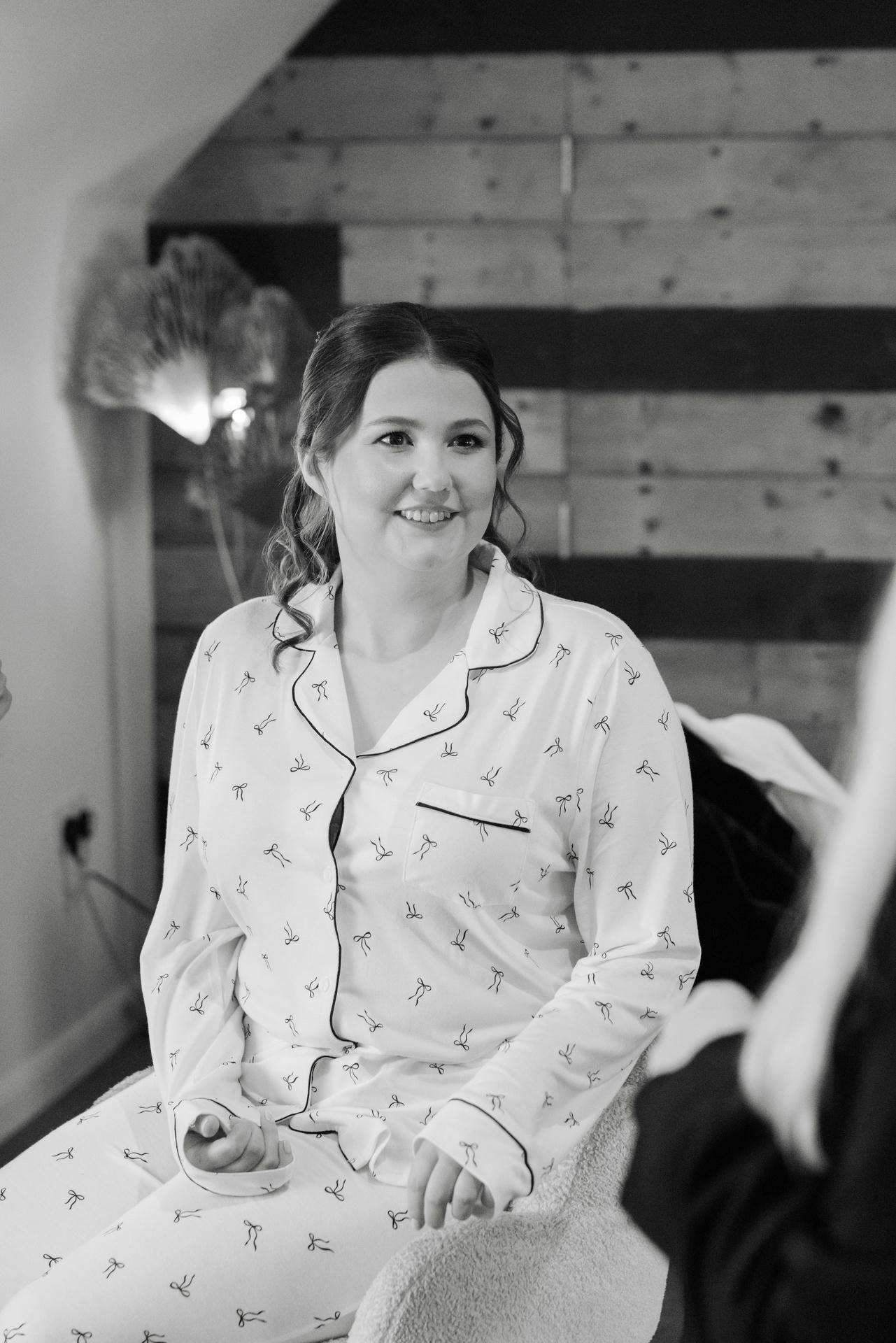 A smiling woman in bow-patterned pajamas sits on a chair in a cozy room with a wooden wall backdrop.