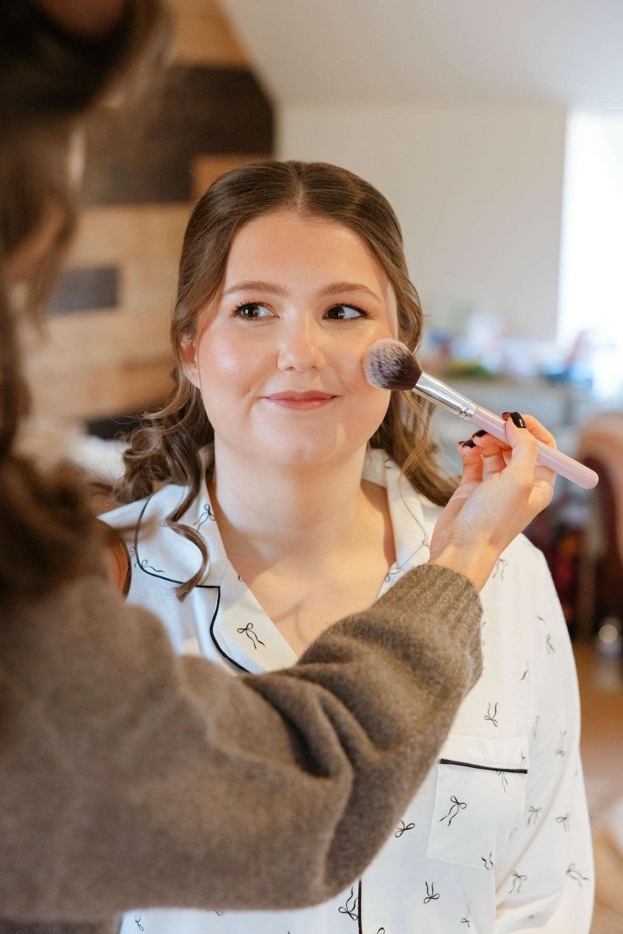 A smiling young woman has blush applied to her cheek by a makeup artist using a fluffy brush.