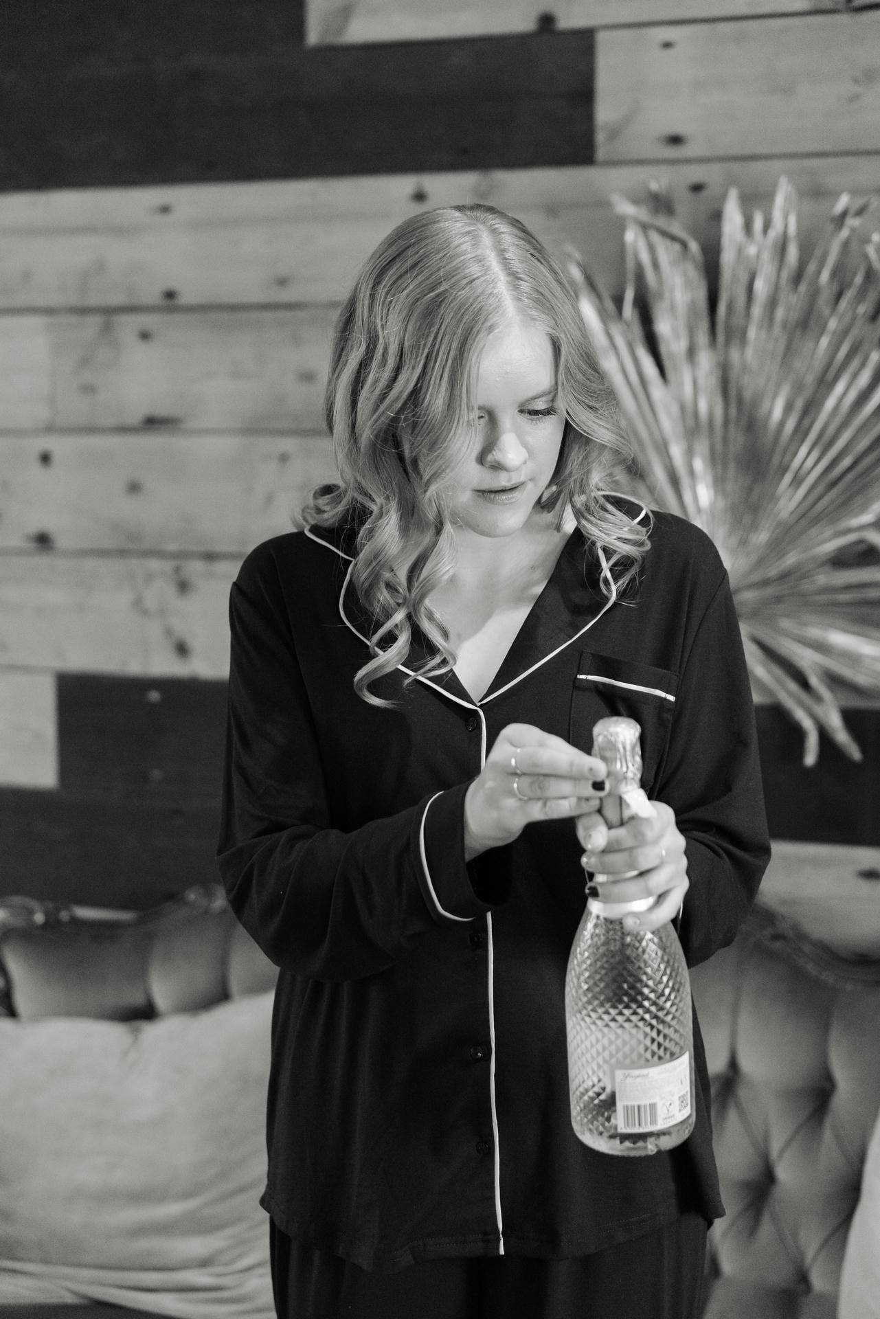 A woman in black pajamas with white piping opens a champagne bottle indoors.