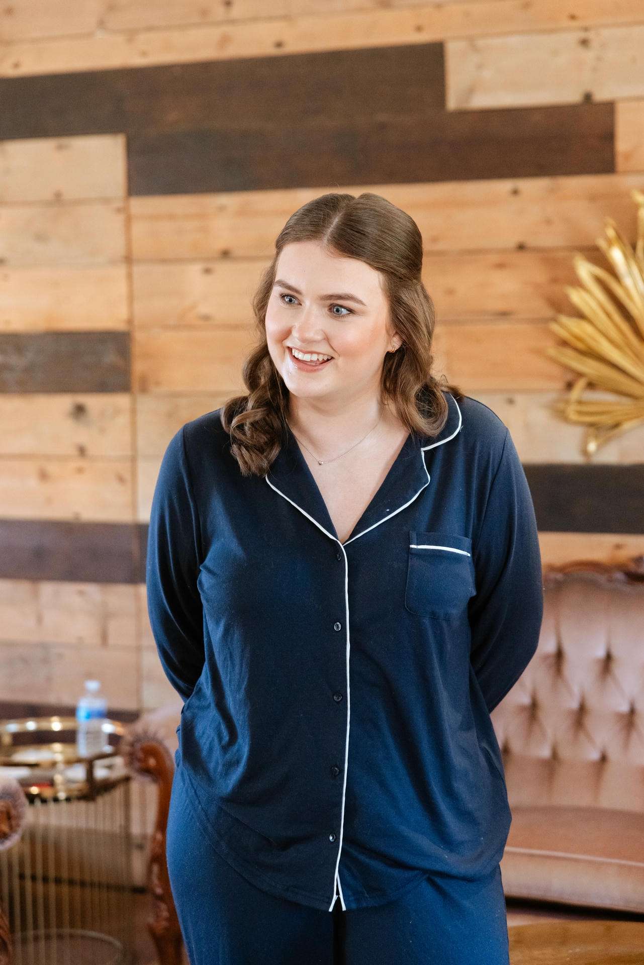 Smiling woman in navy blue pajamas with white piping stands in a wood-paneled room.