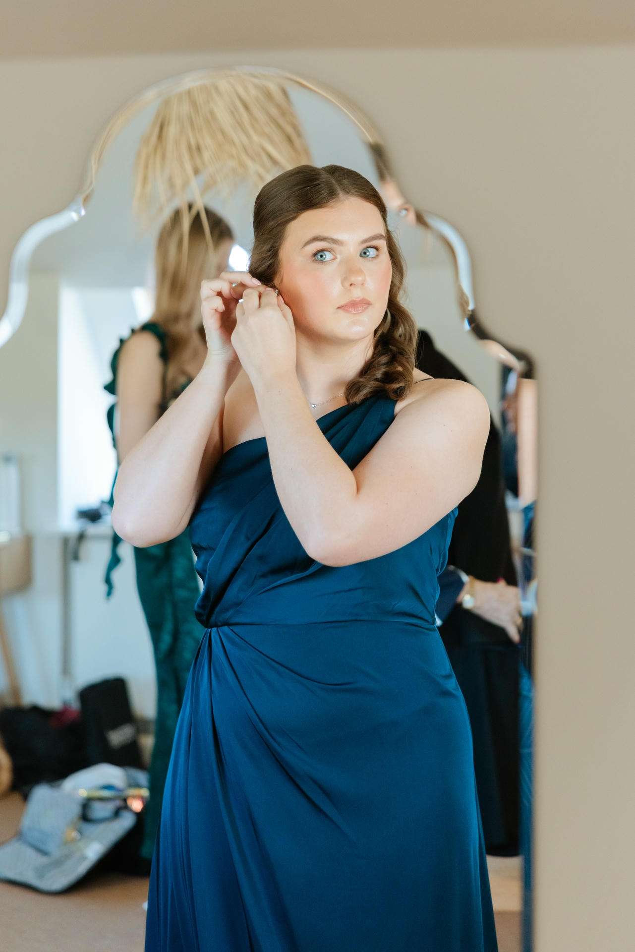 A woman in a teal blue, one-shoulder gown adjusts an earring in front of a large ornate mirror.