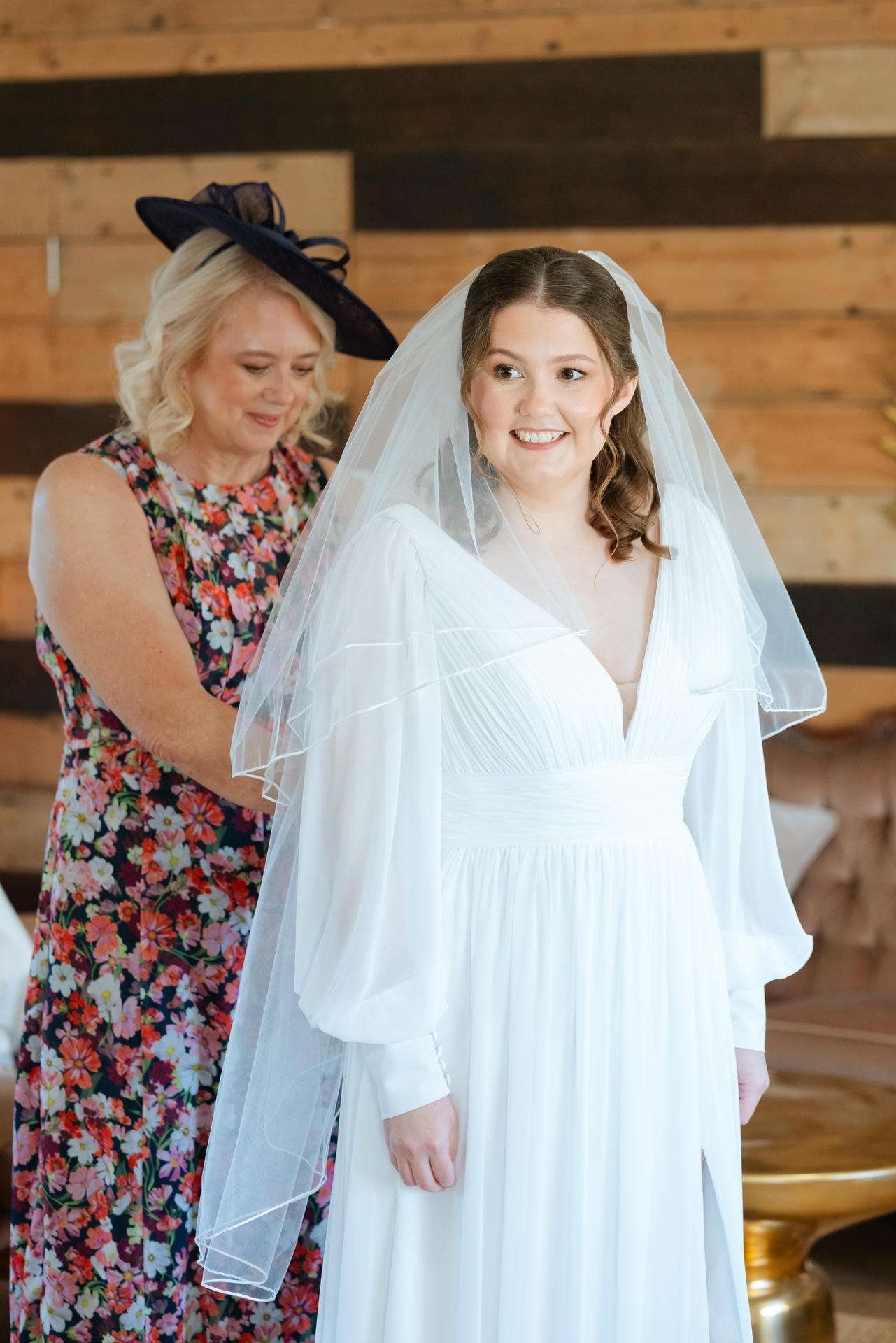 Bride in a white wedding dress with a veil smiles as a woman in a floral dress and a dark hat assists her.