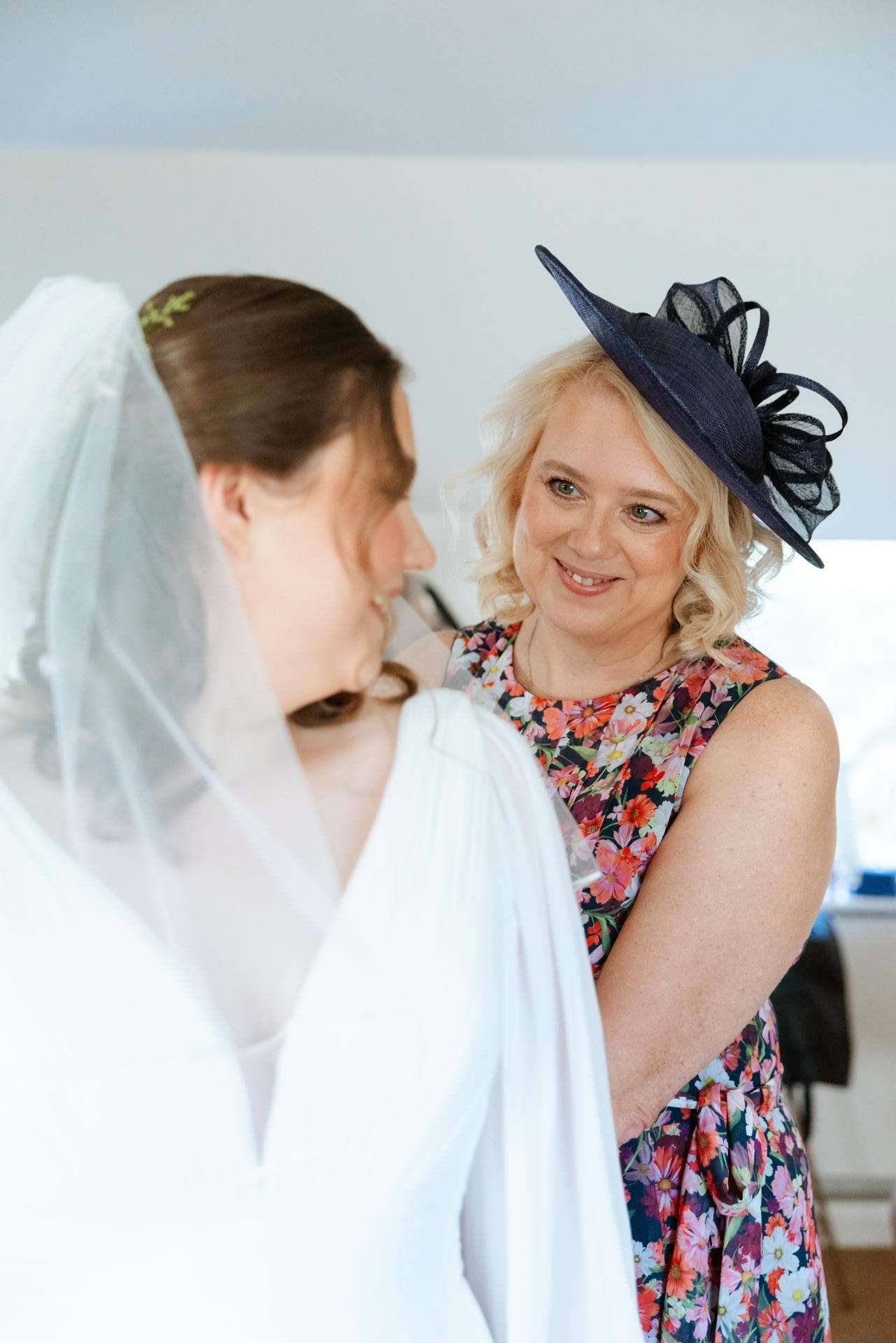 A bride in a white wedding gown and veil smiles at a woman in a colorful floral dress wearing a navy hat.