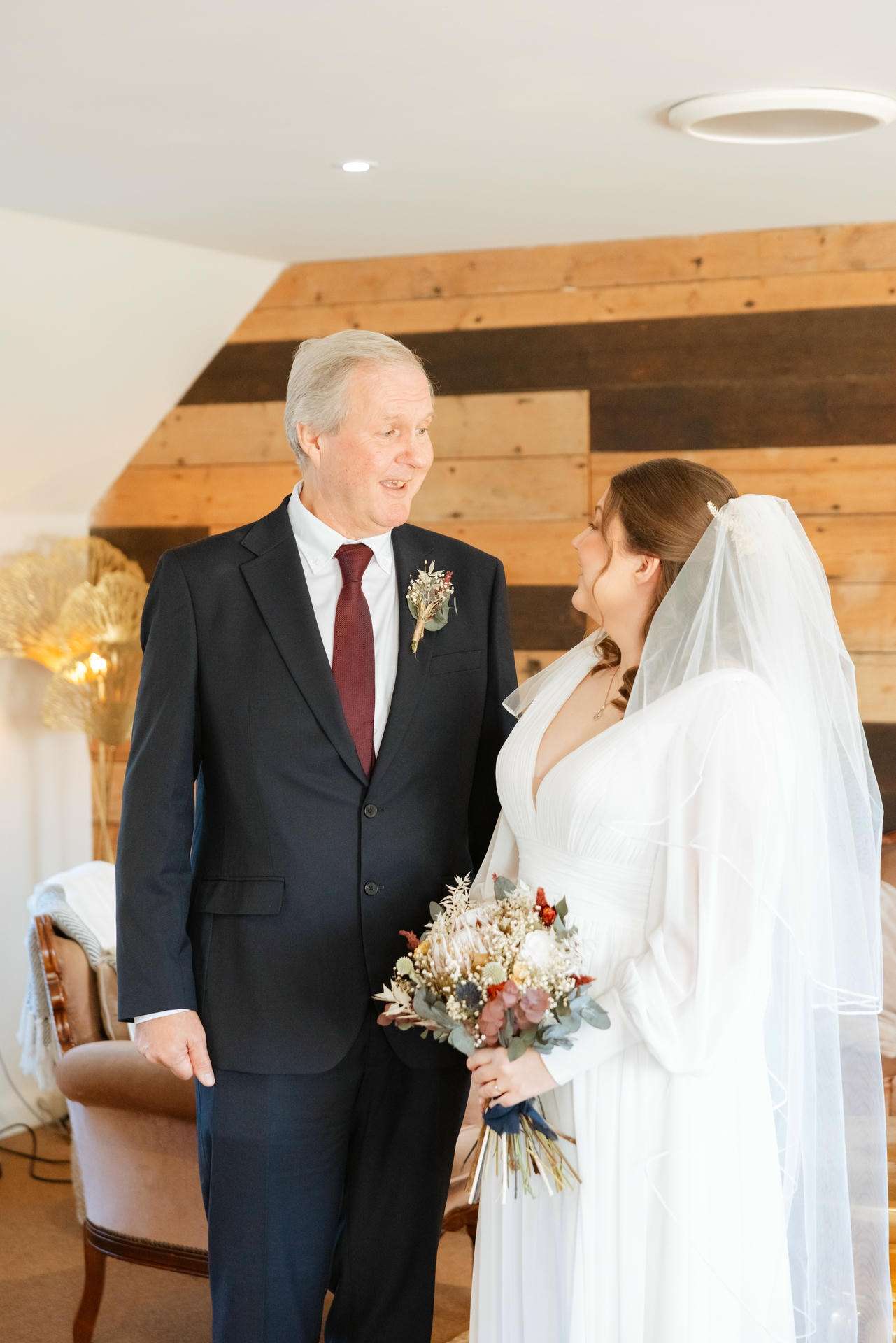 Bride in a white wedding gown with veil holds a bouquet and stands next to a man in a dark suit indoors against a wooden paneled wall.