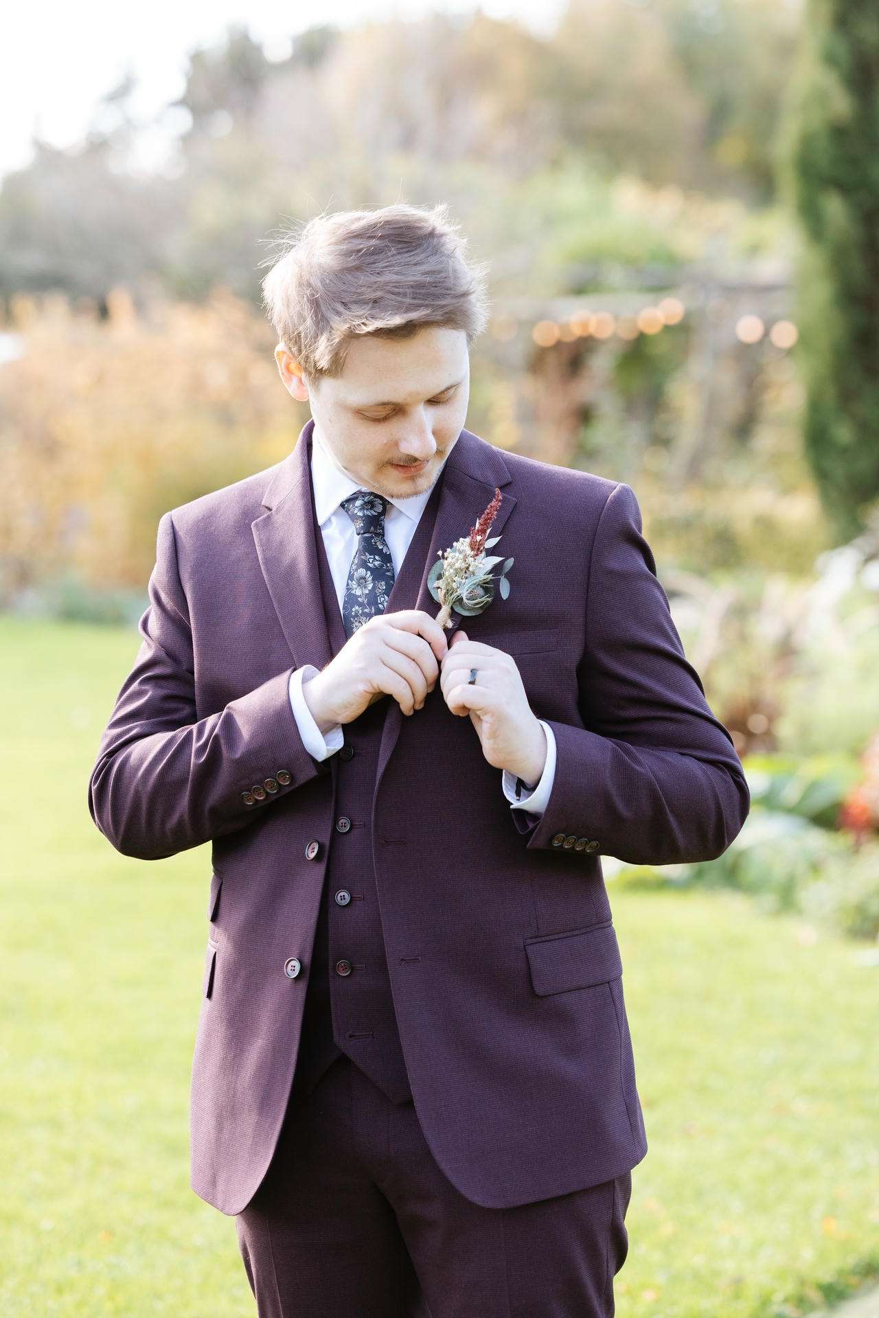 A man in a burgundy suit pins a boutonniere to his lapel in a garden.