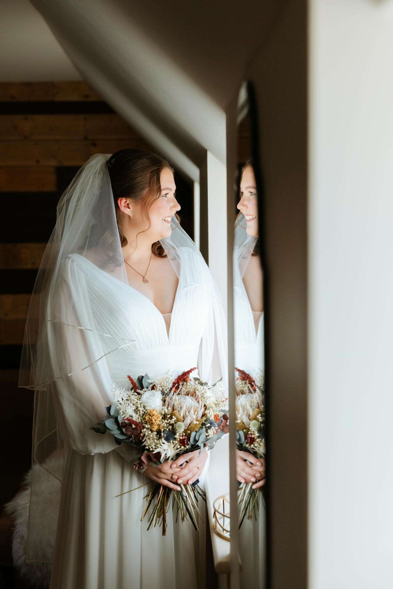 A bride in a white wedding dress and veil holds a dried-flower bouquet and smiles at her reflection in a windowpane.