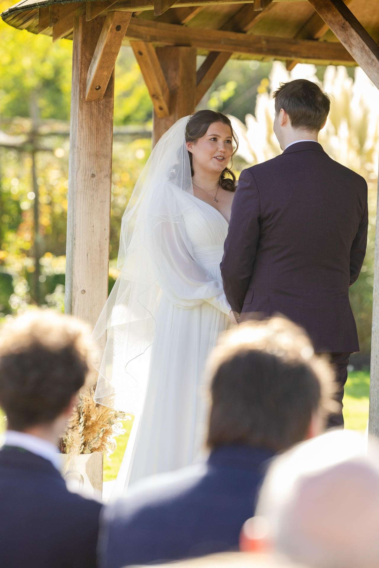 Bride in a white wedding dress and veil speaks with a groom in a dark suit under a wooden gazebo at an outdoor ceremony.