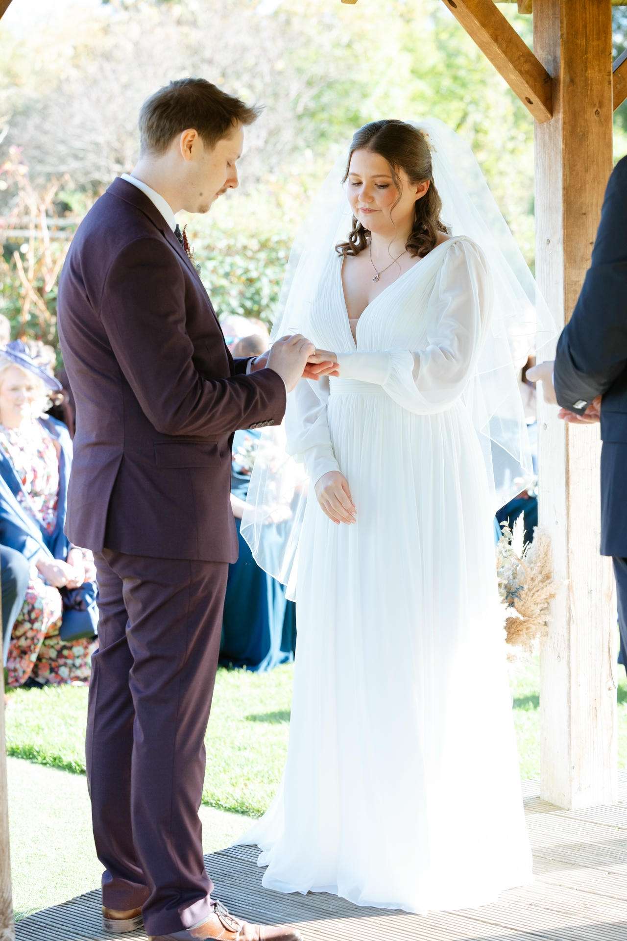 A bride in a white wedding dress and veil exchanges rings with a groom in a dark suit during an outdoor ceremony under a wooden arch.
