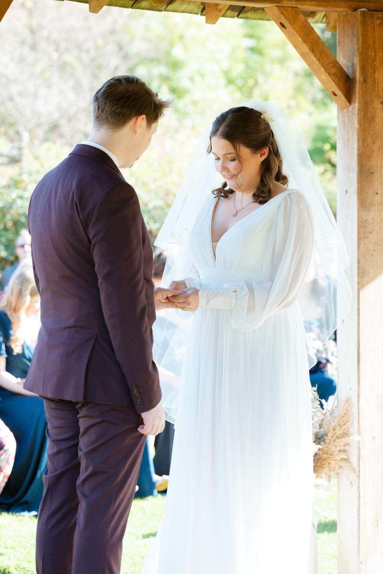 Bride placing a ring on the groom's finger during an outdoor wedding under a wooden gazebo.