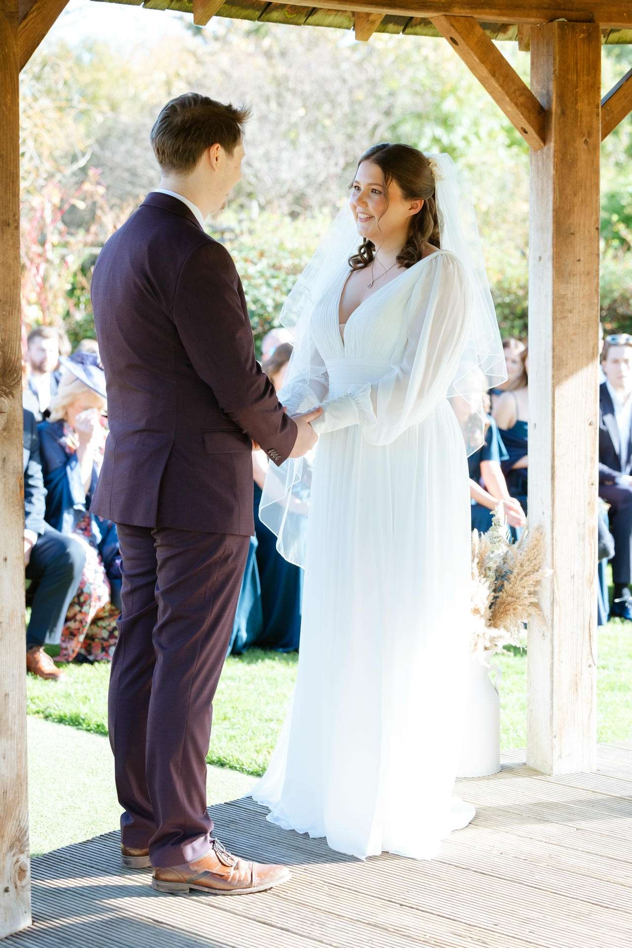 Bride in a white gown and veil holds hands with a groom in a burgundy suit under a wooden gazebo at an outdoor wedding.
