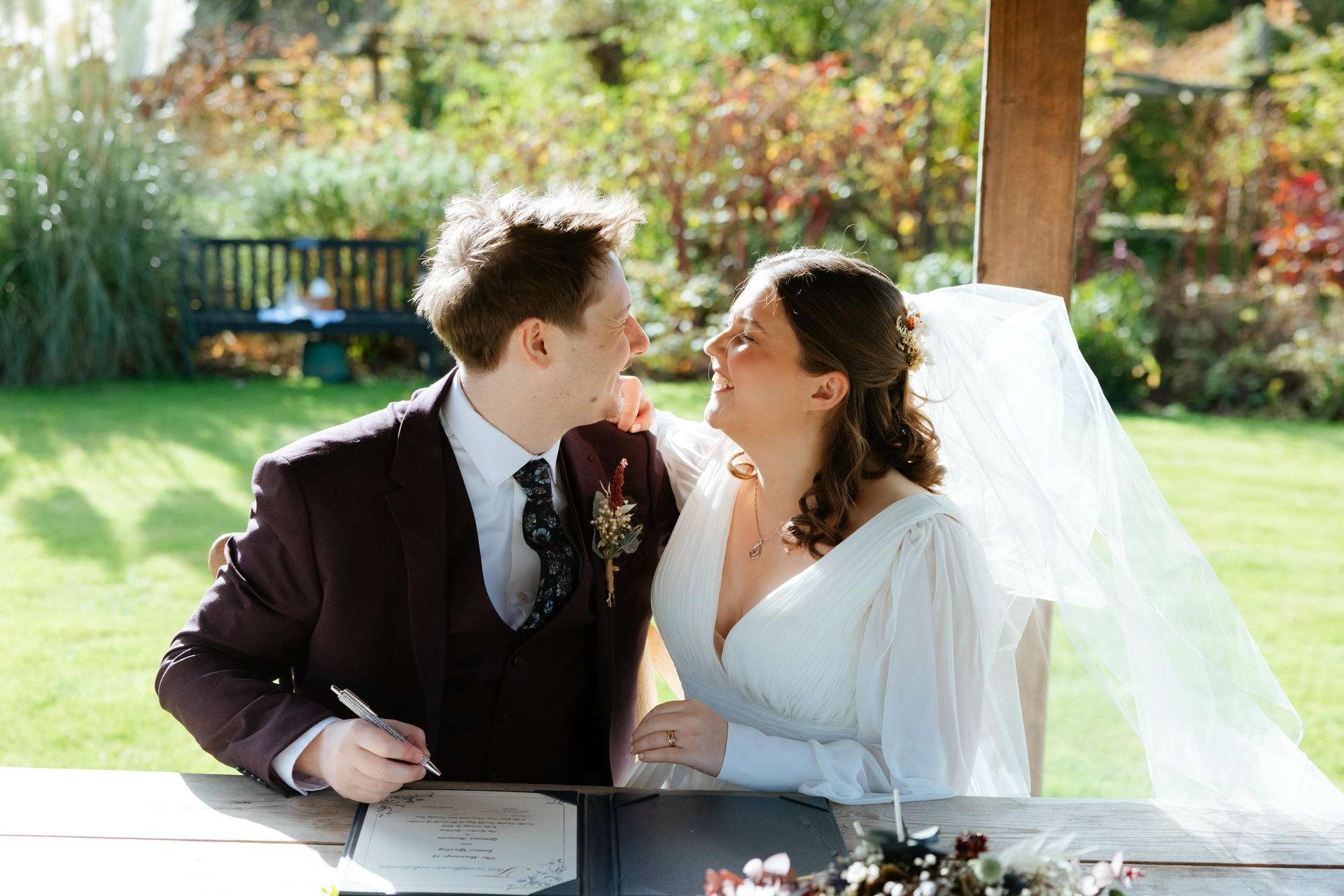 A bride and groom in wedding attire sign documents at an outdoor table, smiling at each other.