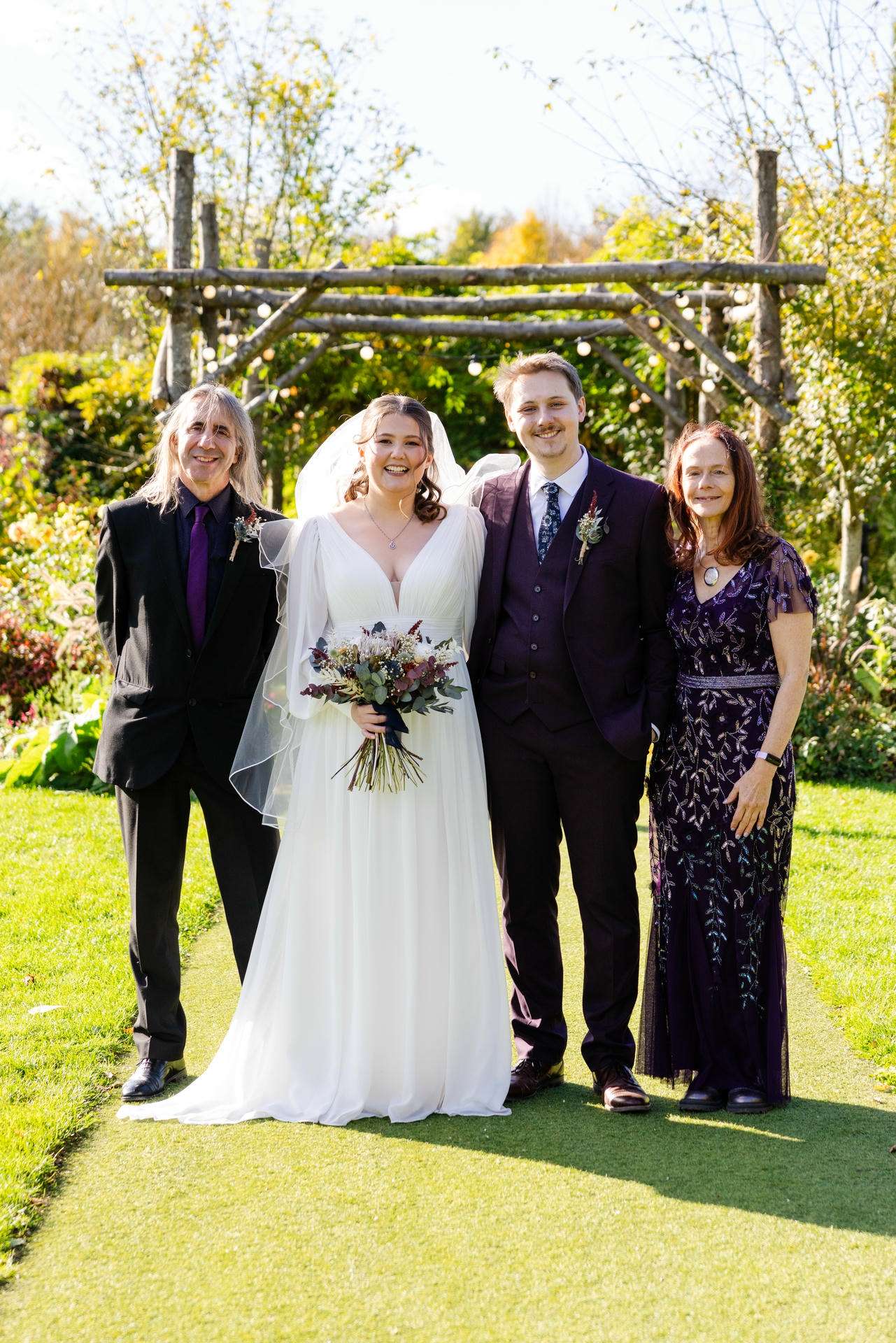Bride in a white wedding gown and veil, holding a bouquet, stands with a groom in a burgundy suit and two guests outdoors under a wooden garden arch.