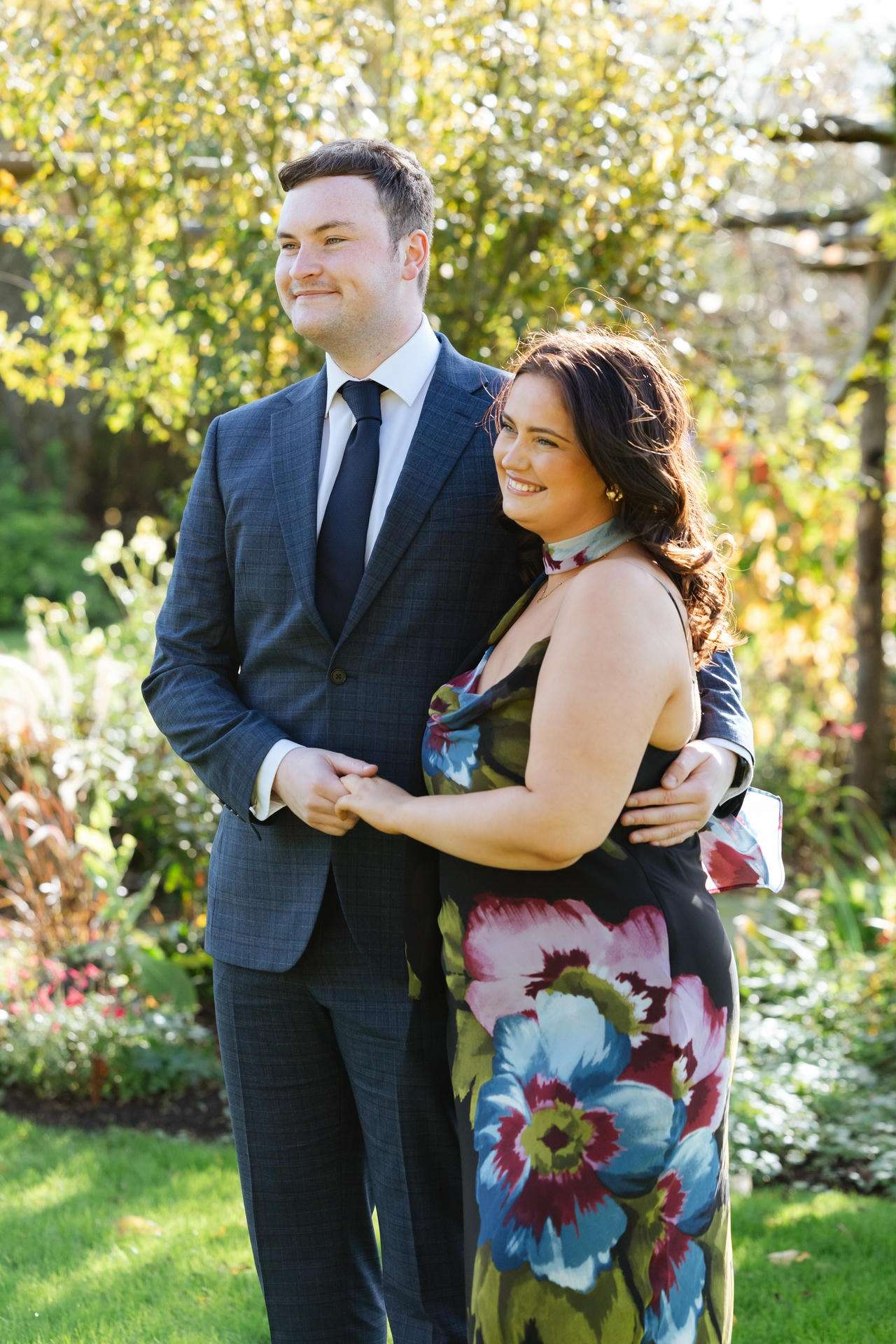 A couple stands in a sunlit garden, the man wearing a blue suit and tie and the woman in a colorful floral dress, smiling while holding hands.
