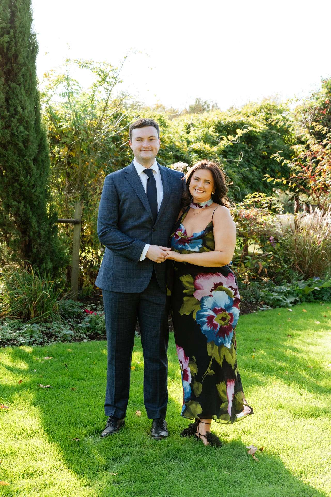 A smiling couple stands in a sunlit garden, the man in a blue suit and the woman in a colorful floral dress, holding hands.