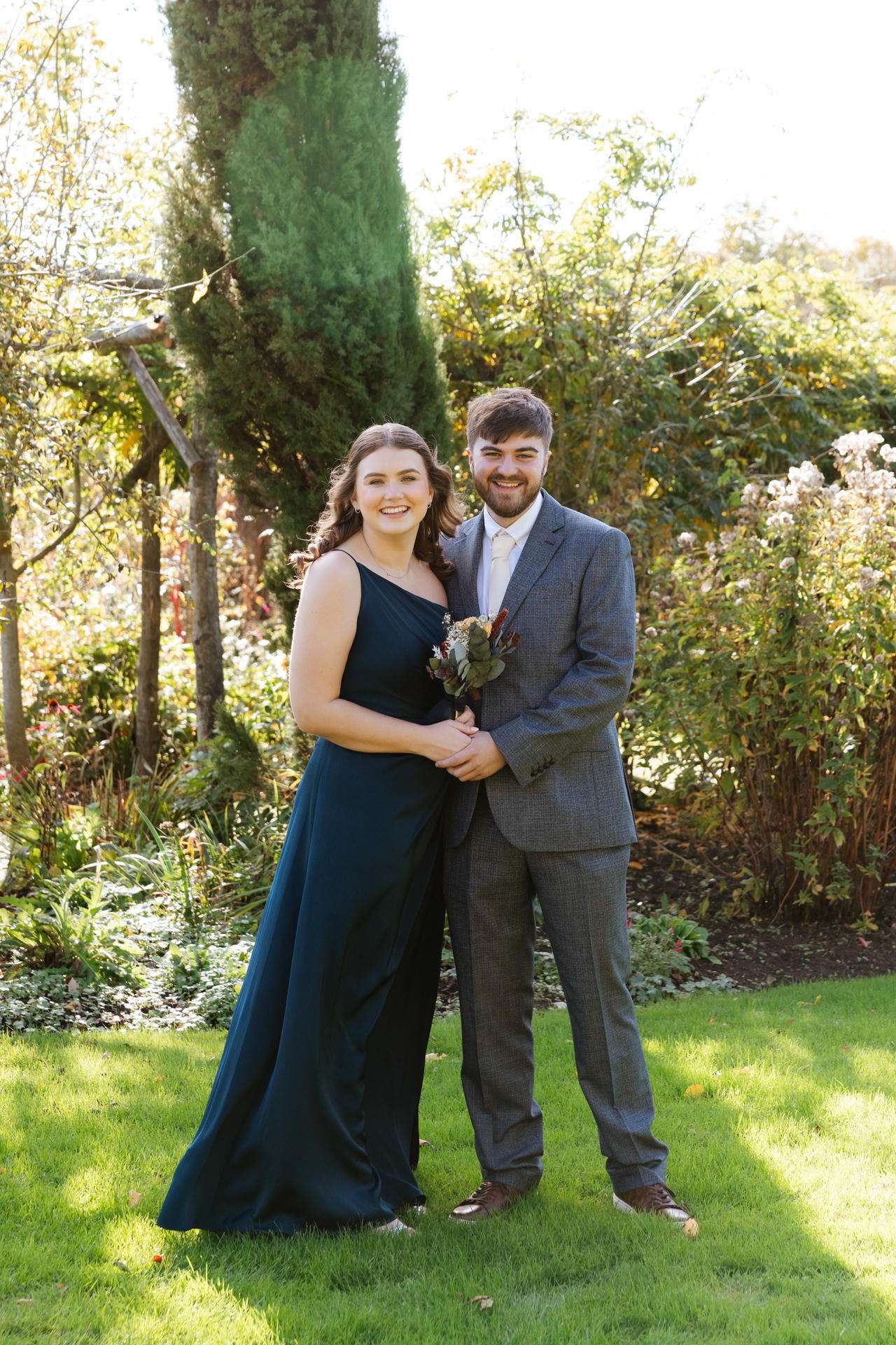 A smiling couple stands in a sunlit garden, the woman in a dark teal one-shoulder gown and the man in a gray suit, holding a small bouquet.
