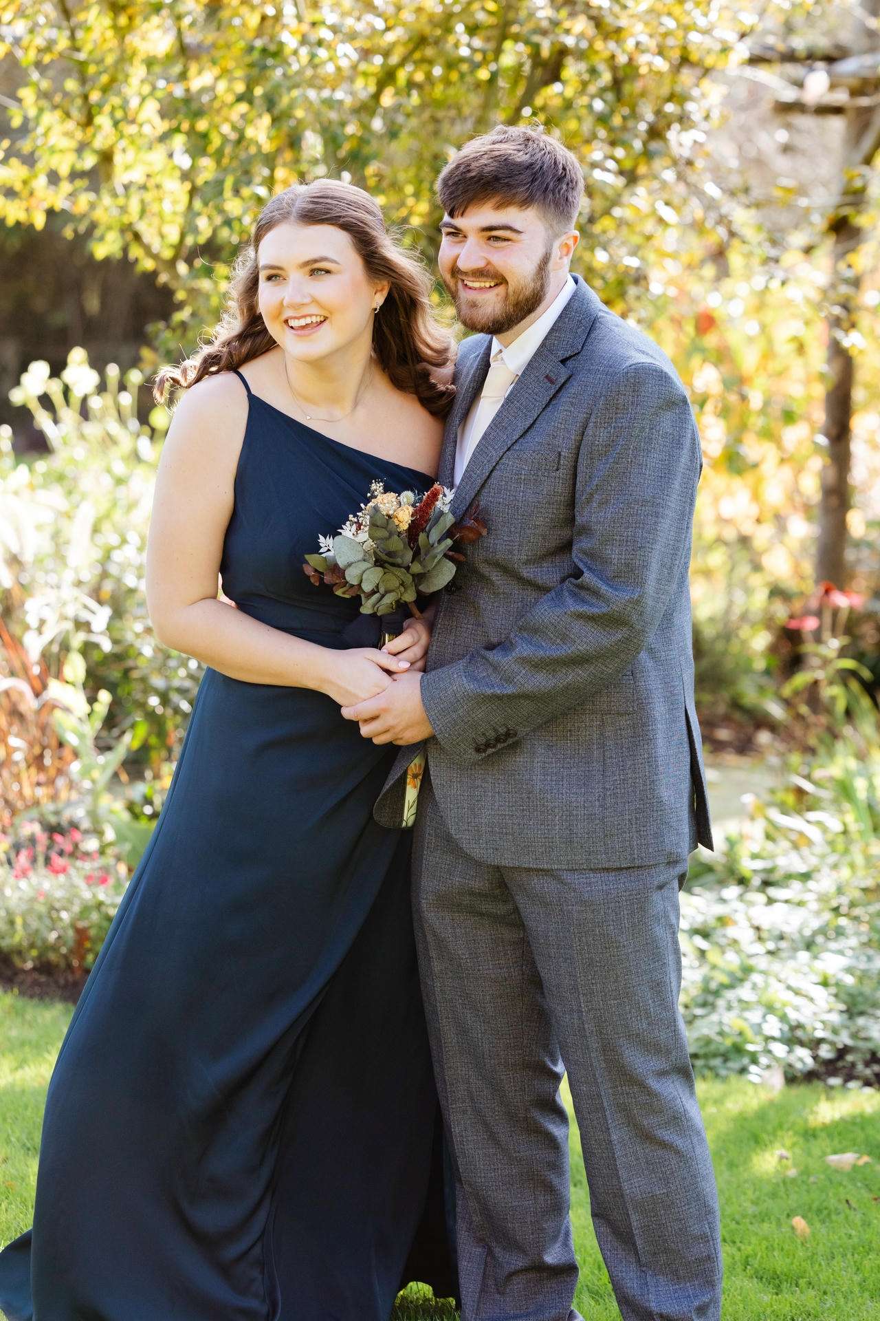 A couple posing together in a sunlit garden, the woman in a navy blue one-shoulder dress holding a small bouquet and the man in a gray suit.