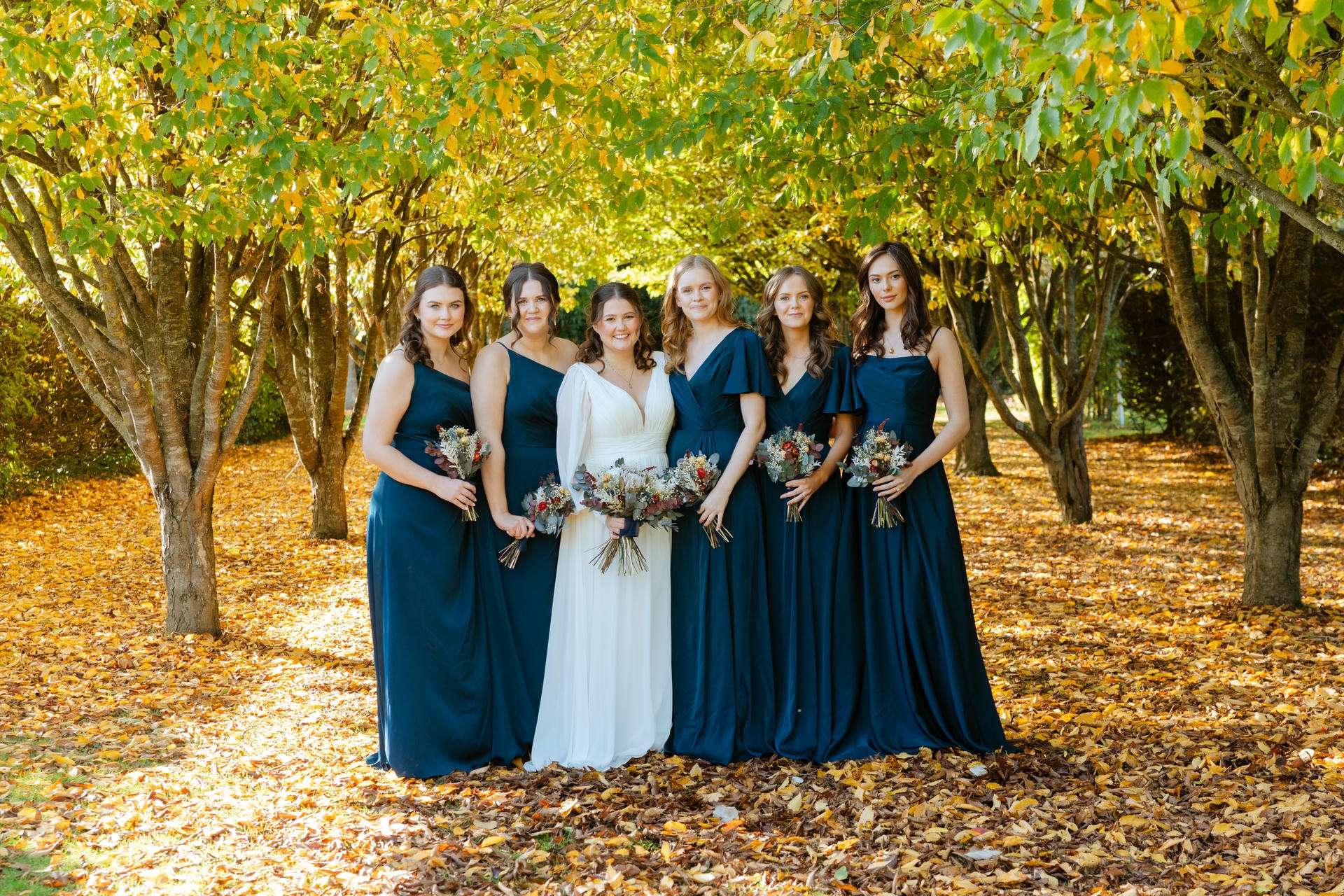Bride in a white gown stands with six bridesmaids in teal dresses on an autumn leaf-covered path under yellow-green trees.