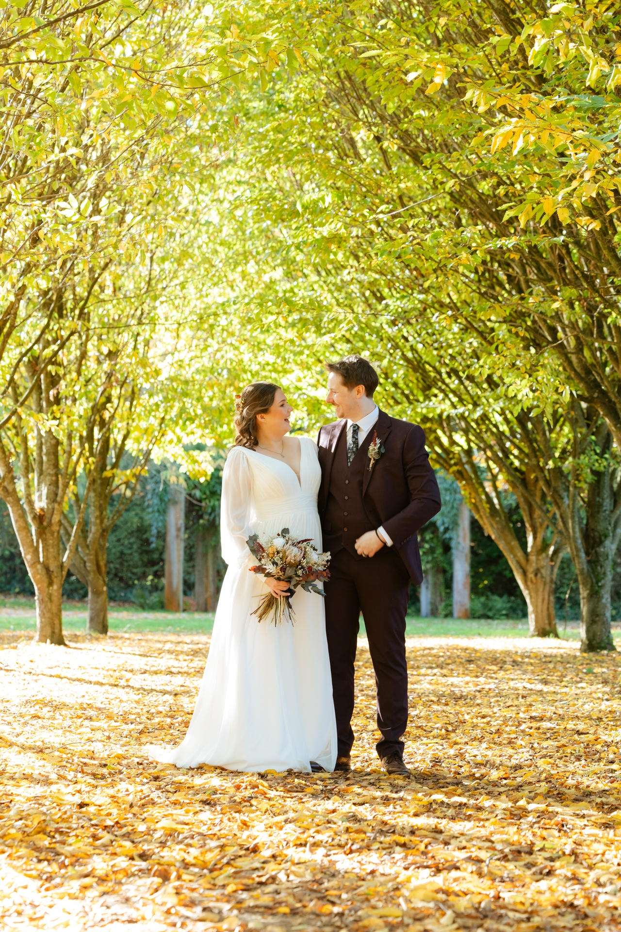 Bride in a white wedding dress and groom in a dark suit walk arm in arm along a leaf-strewn path under sunlit autumn trees.
