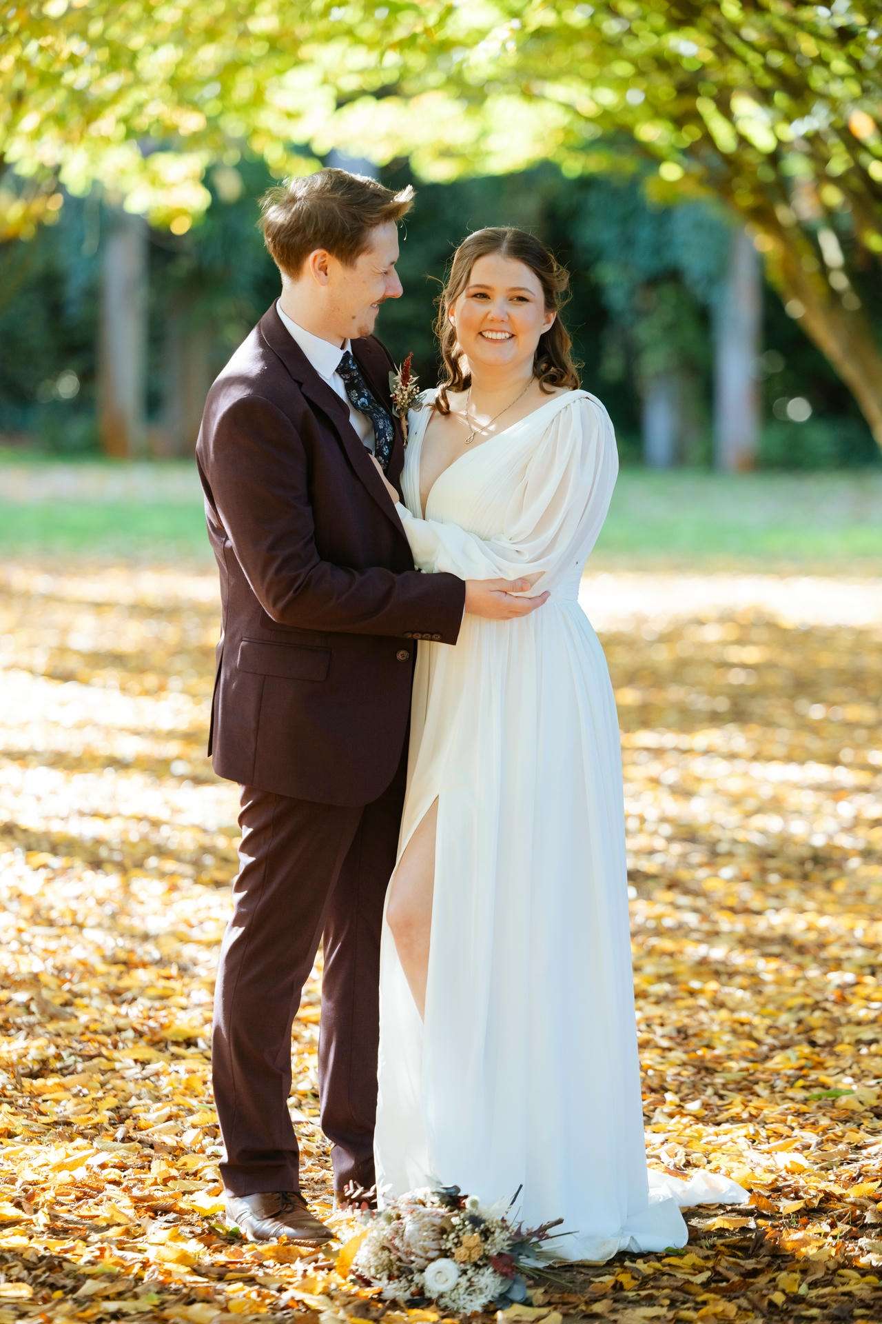 A bride in a white gown and a groom in a maroon suit stand outdoors on a leaf-strewn path, smiling at each other.