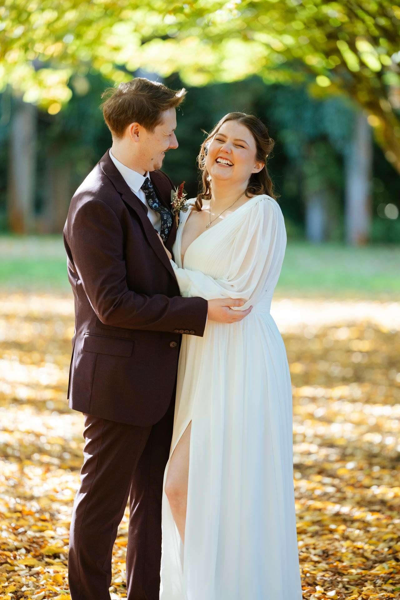 A couple in formal attire hugs and laughs in a sunlit park with autumn leaves on the ground.