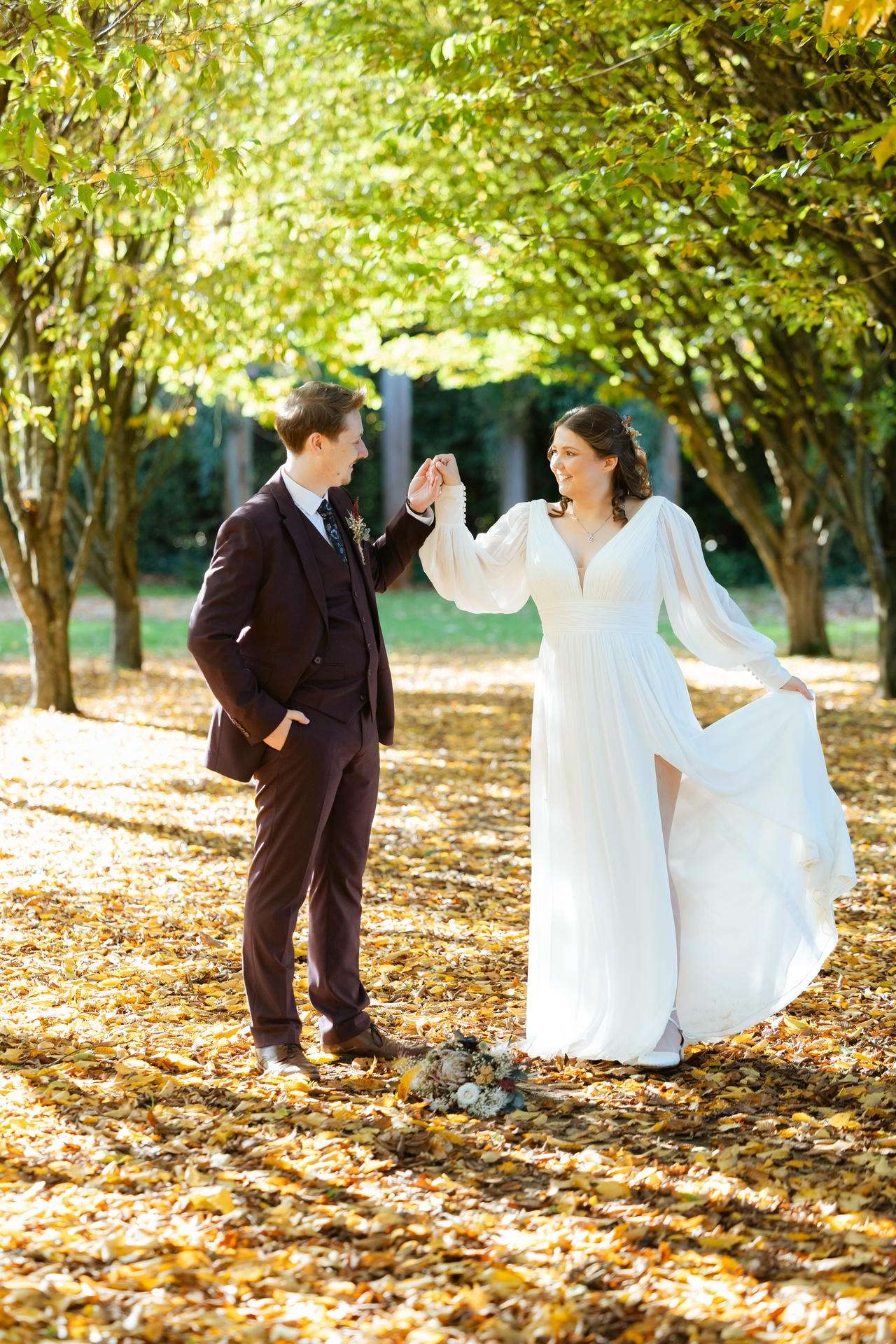 Bride in a white gown and groom in a maroon suit hold hands and smile on a leaf-covered path beneath a canopy of trees.