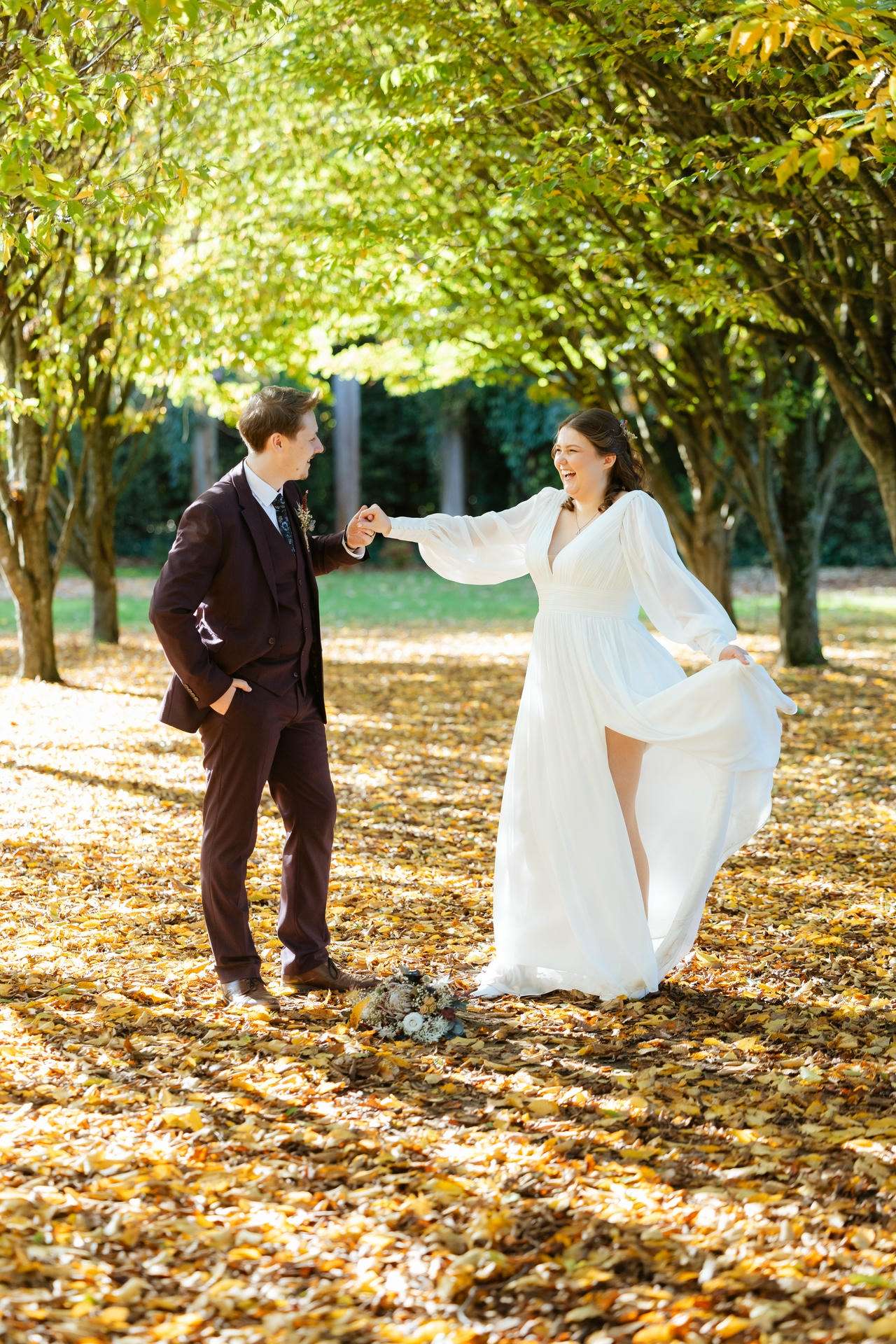 A bride in a flowing white gown and a groom in a burgundy suit hold hands and smile while walking through a sunlit autumn park covered in fallen leaves.
