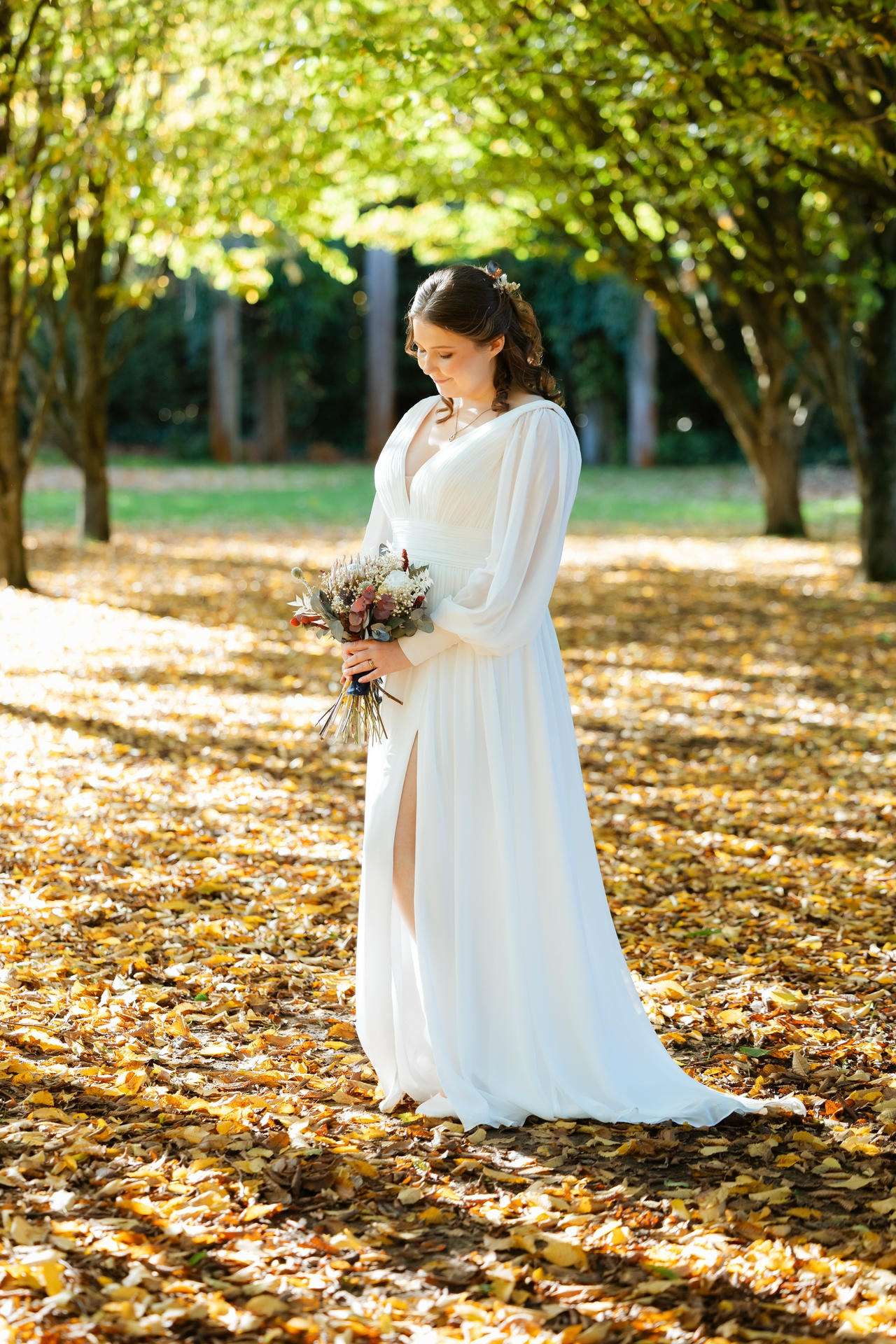 Bride in a flowing white gown holding a bouquet stands in a sunlit park covered with autumn leaves.