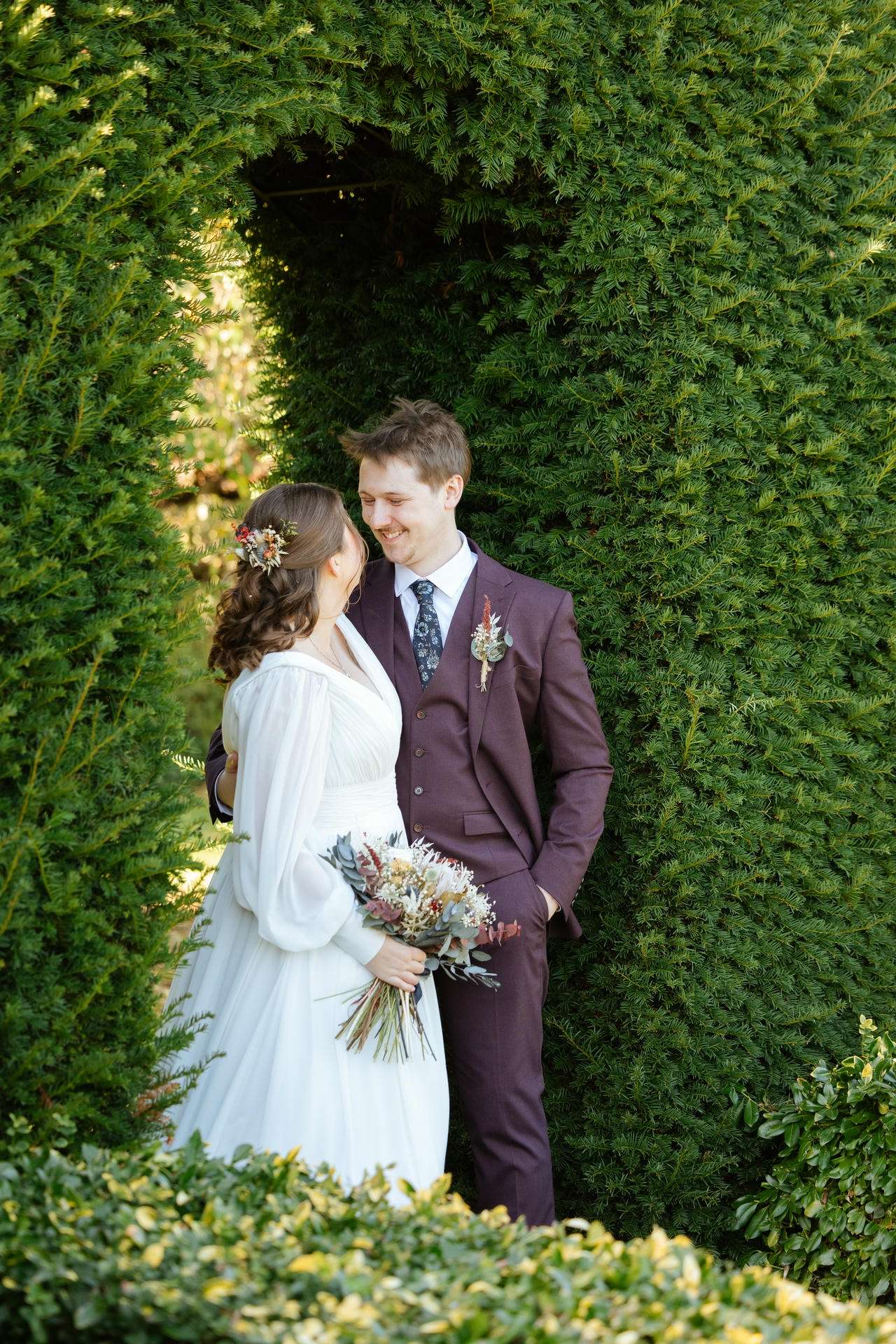 A bride in a white dress and a groom in a burgundy suit smile at each other under a dense green hedge arch, the bride holding a dried-flower bouquet.