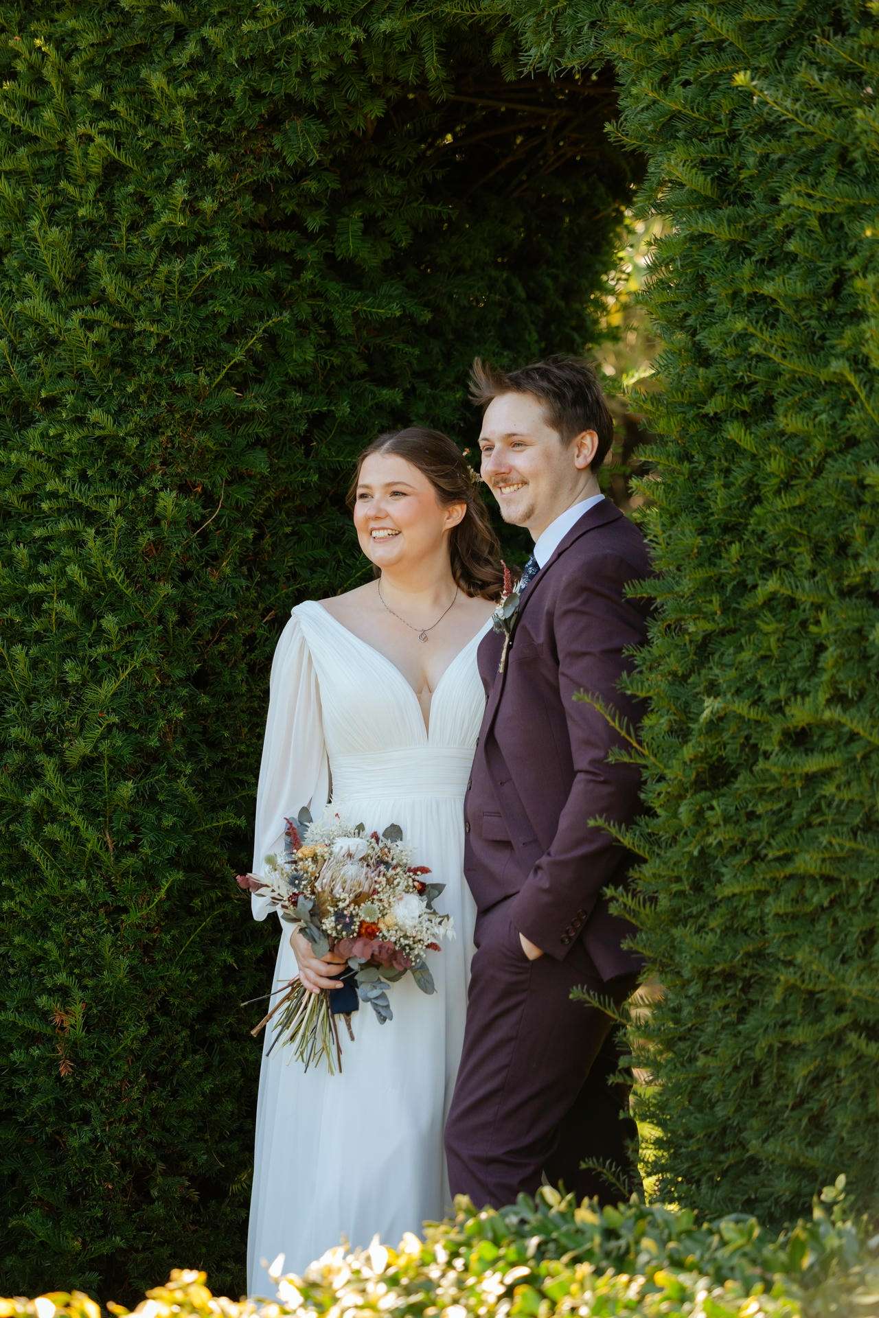 Bride in a white gown and groom in a burgundy suit stand together and smile beneath a lush green hedge arch, the bride holding a dried-flower bouquet.