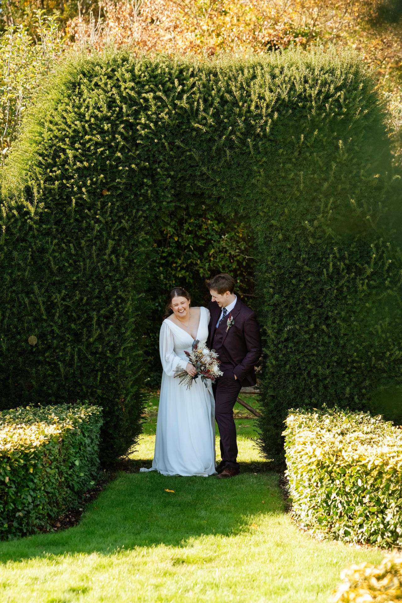 A bride in a pale blue wedding dress holds a bouquet as she and a groom in a dark suit stand together beneath a tall hedge arch in a sunlit garden.
