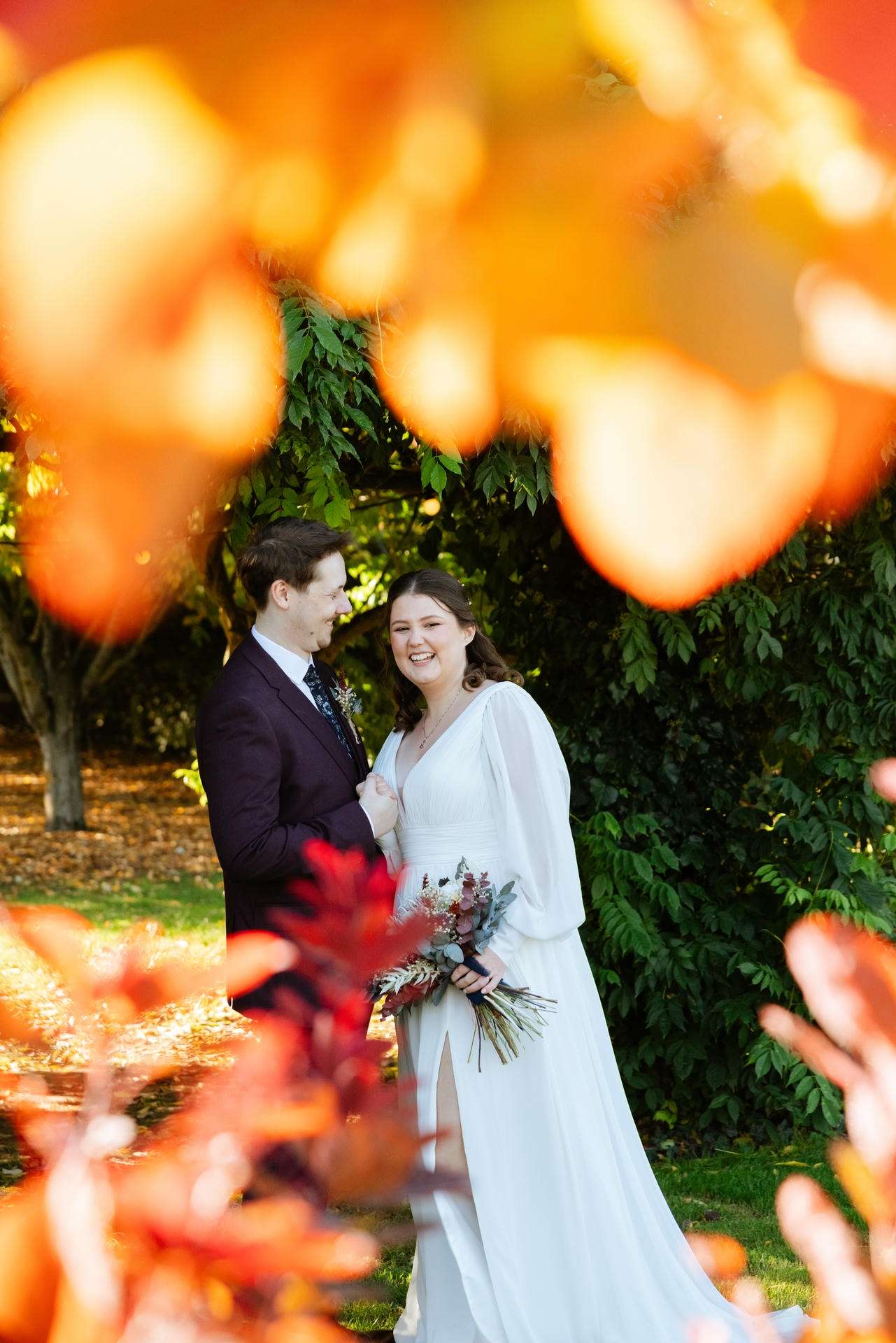 A bride in a white wedding dress holding a bouquet stands with a groom in a dark suit in a sunlit garden framed by vibrant autumn leaves.