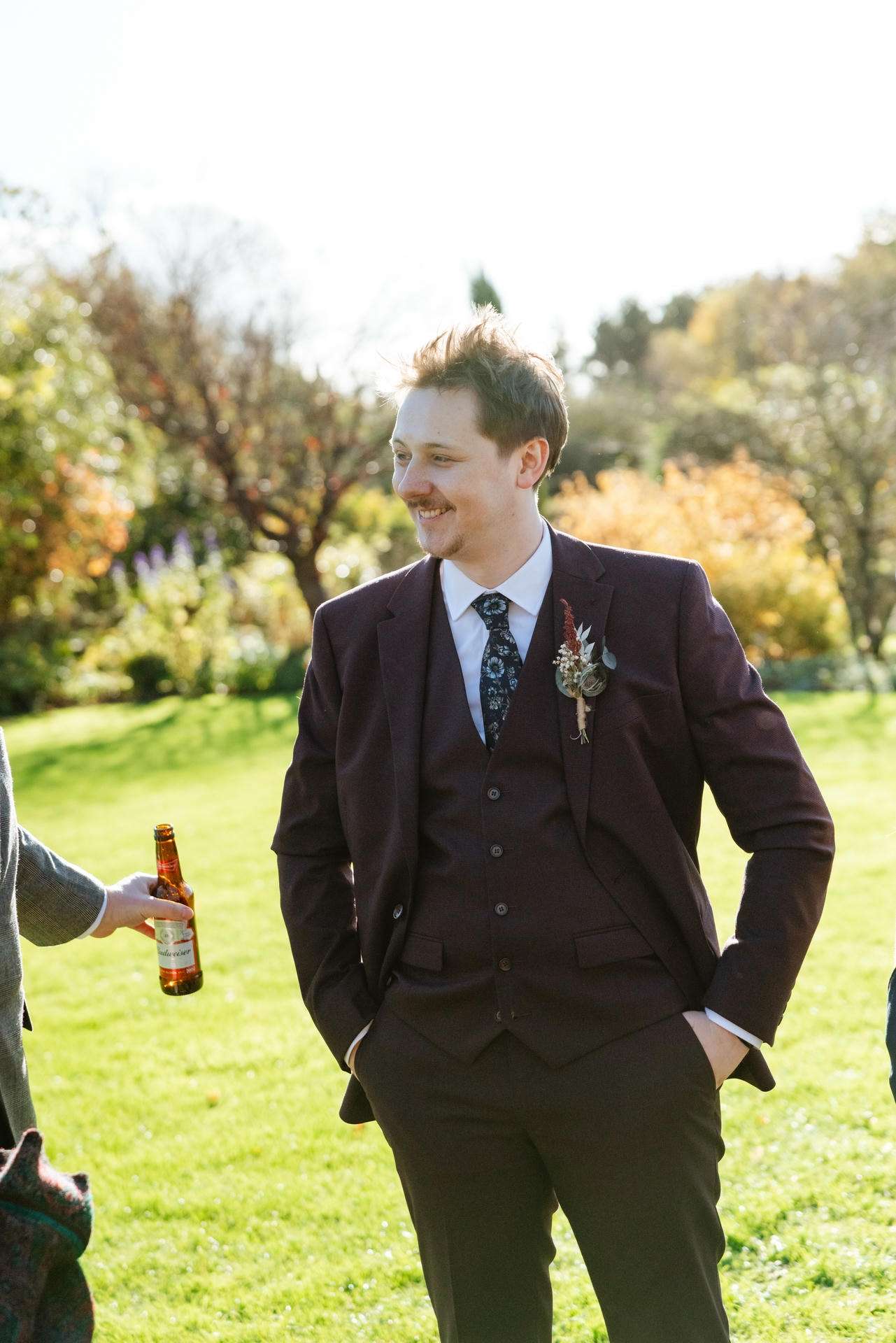 A man in a dark suit with a vest and floral boutonniere stands in a sunlit park, smiling.