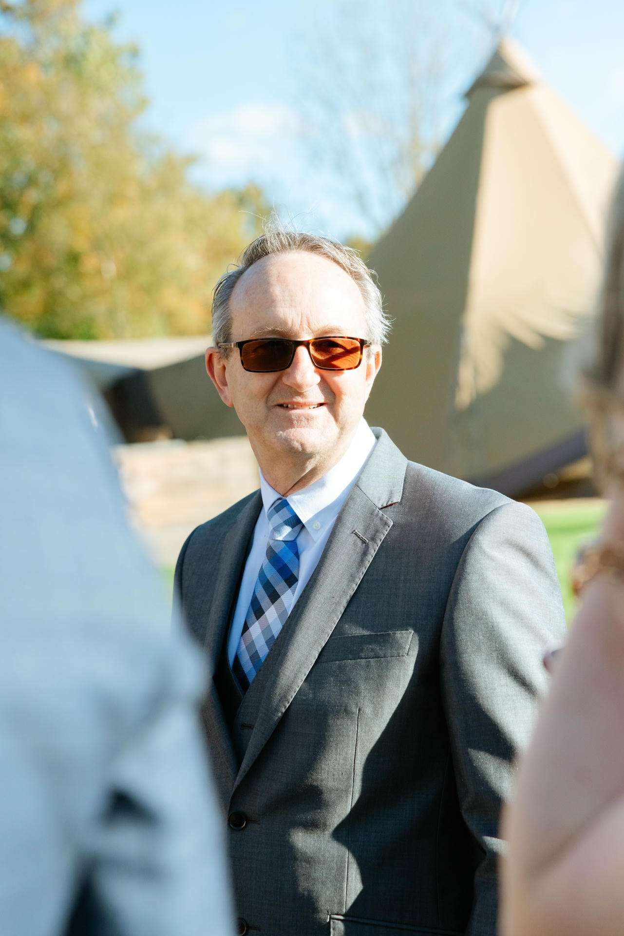 A smiling man in a gray suit and blue striped tie wearing brown sunglasses stands outdoors with tents in the background.