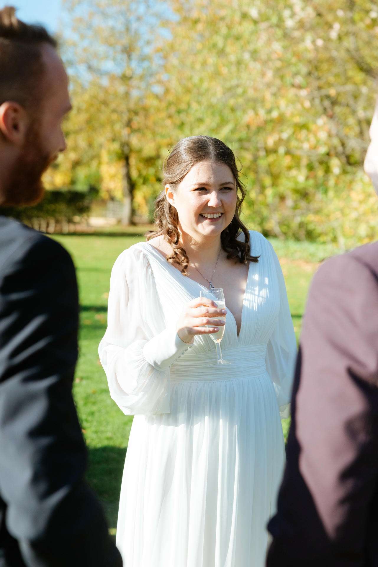Smiling woman in a white dress holding a champagne flute at an outdoor autumn gathering.