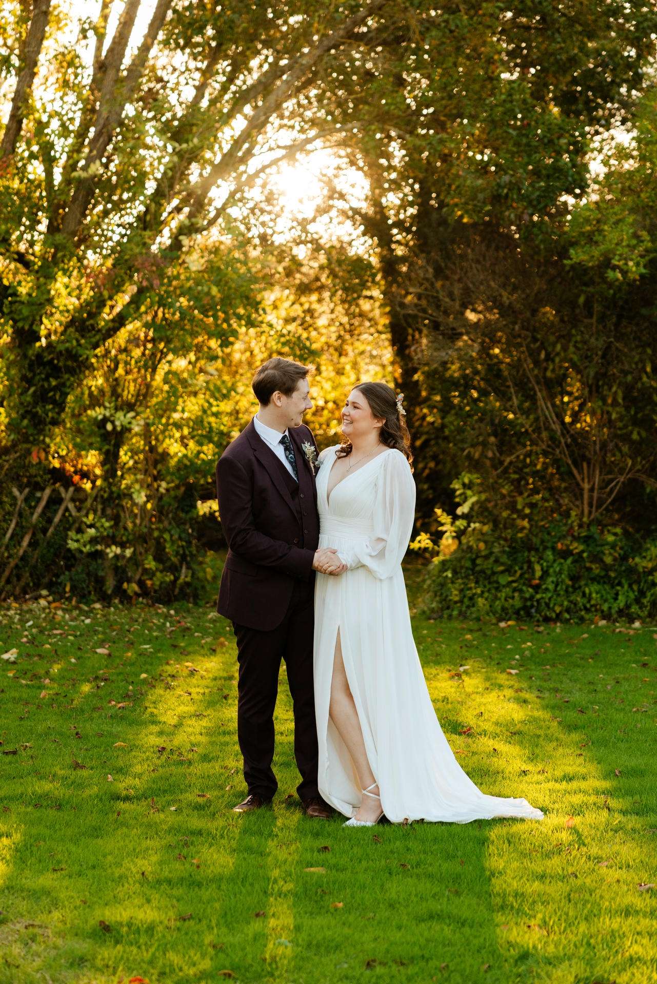 A bride in a white gown with a slit and a groom in a dark suit stand hand in hand on a sunlit lawn, smiling at each other with autumn trees in the background at The Gardens Wedding venue