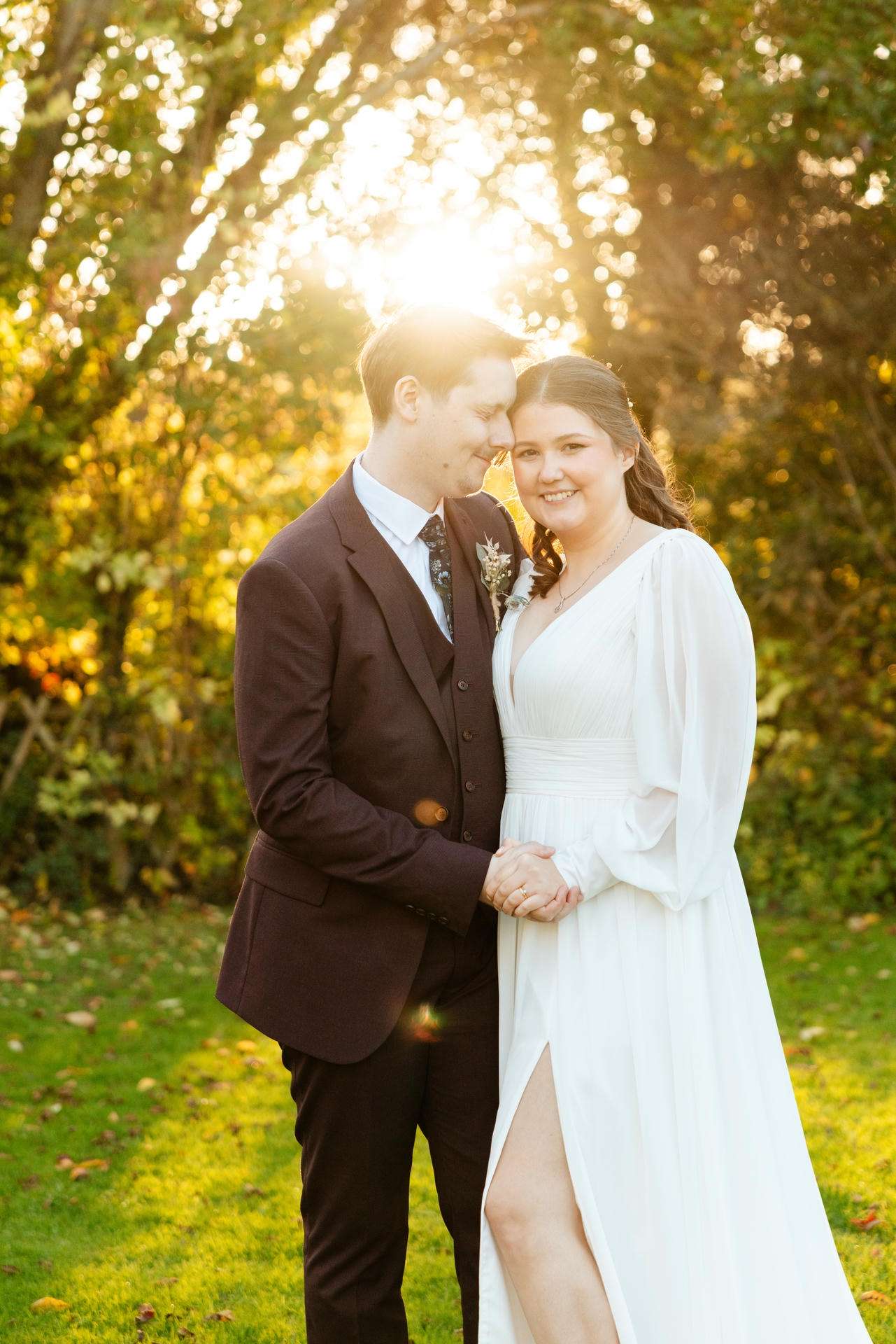 A bride and groom stand hand-in-hand in a sunlit outdoor setting, smiling at The Gardens Wedding venue