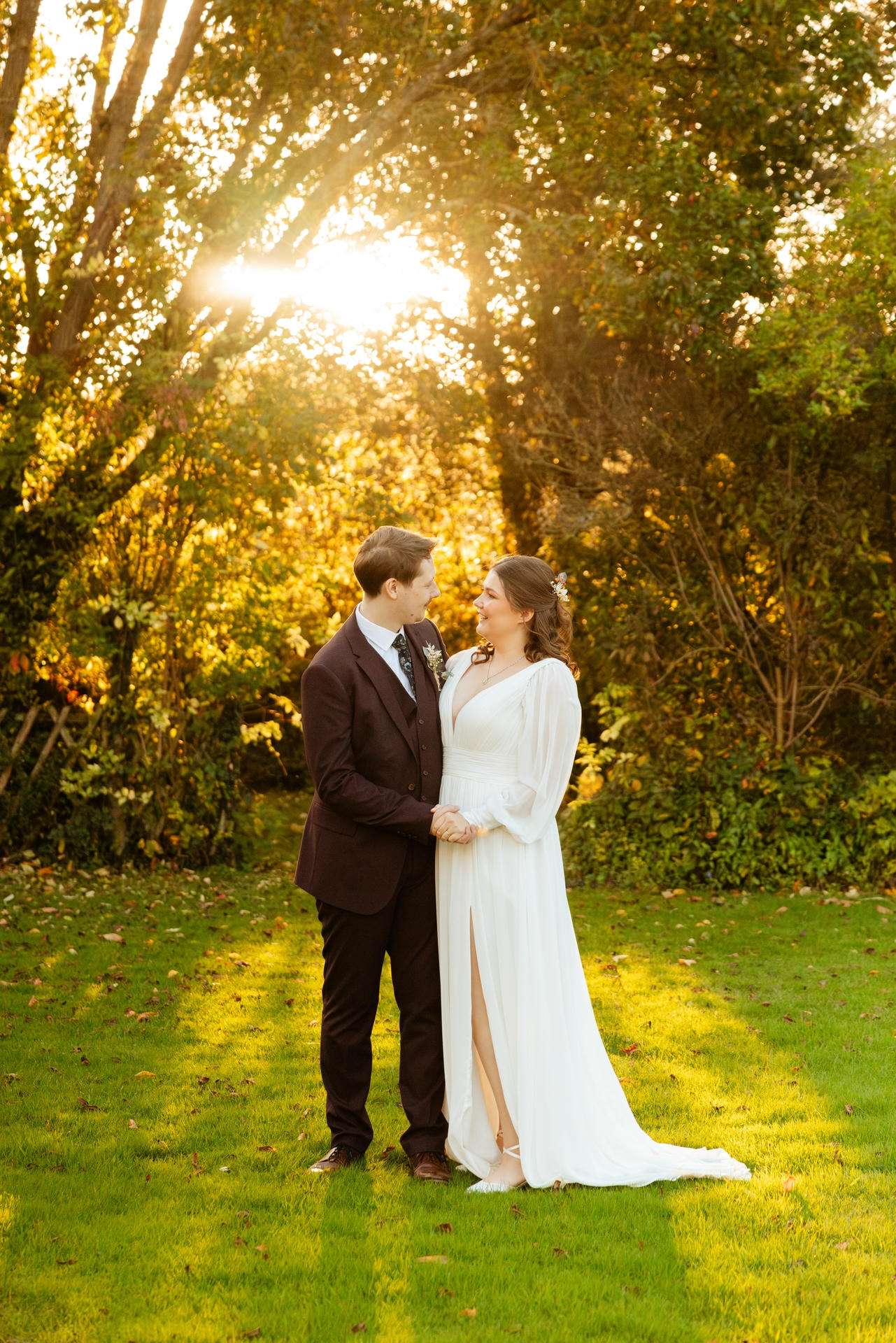 A bride in a white wedding gown and a groom in a dark suit stand hand in hand, smiling at each other in a sunlit autumn garden at The Gardens Wedding venue