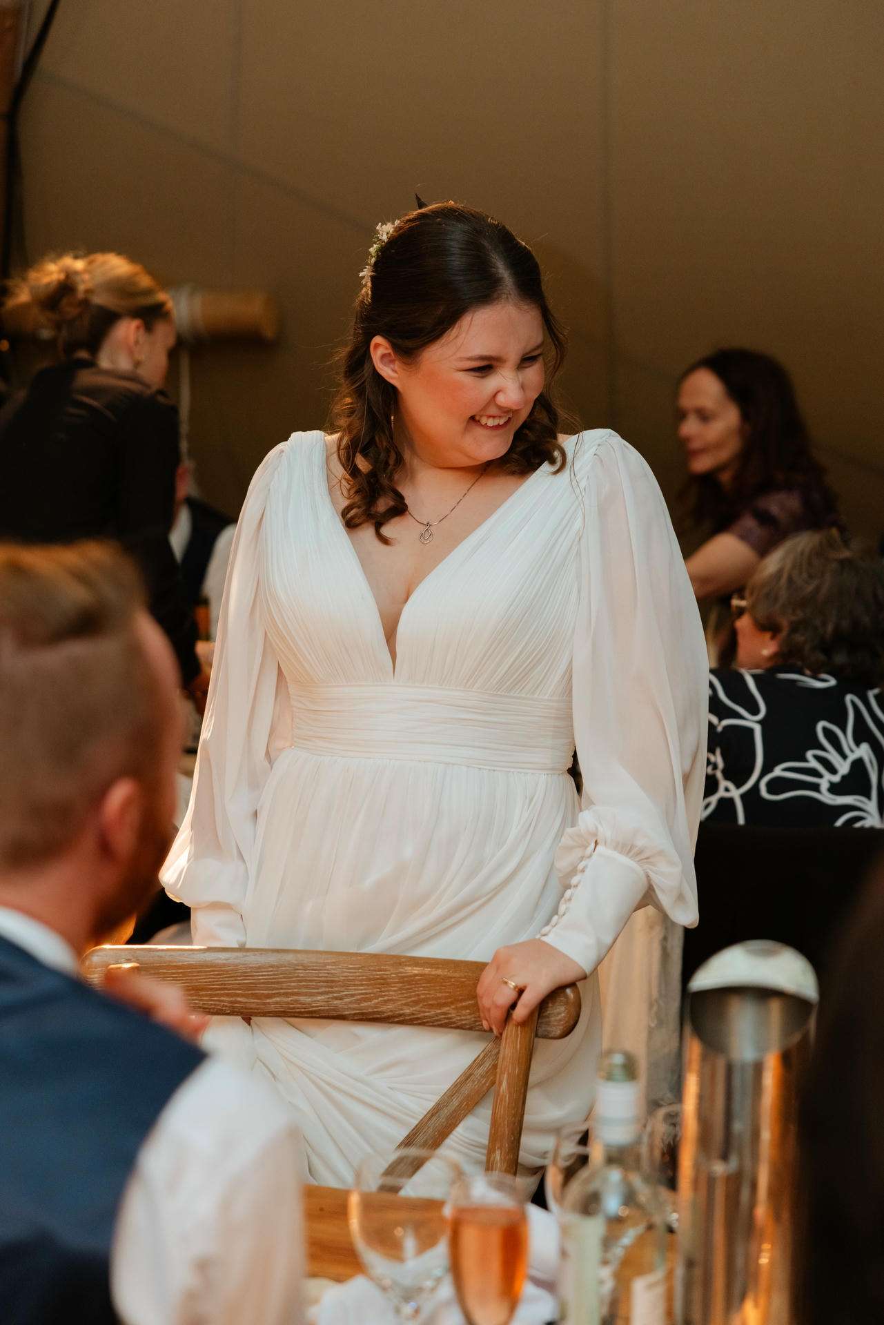 Smiling bride in a white V-neck gown with long sleeves at a wedding reception.