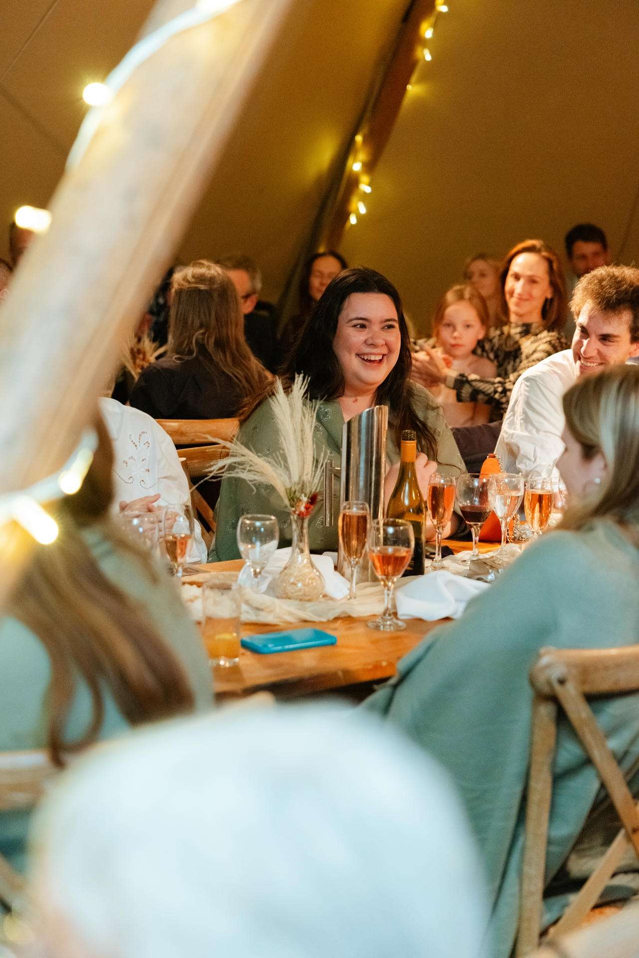 A group of friends sits around a wooden table inside a warmly lit tent, smiling and enjoying wine glasses and a decorative centerpiece.