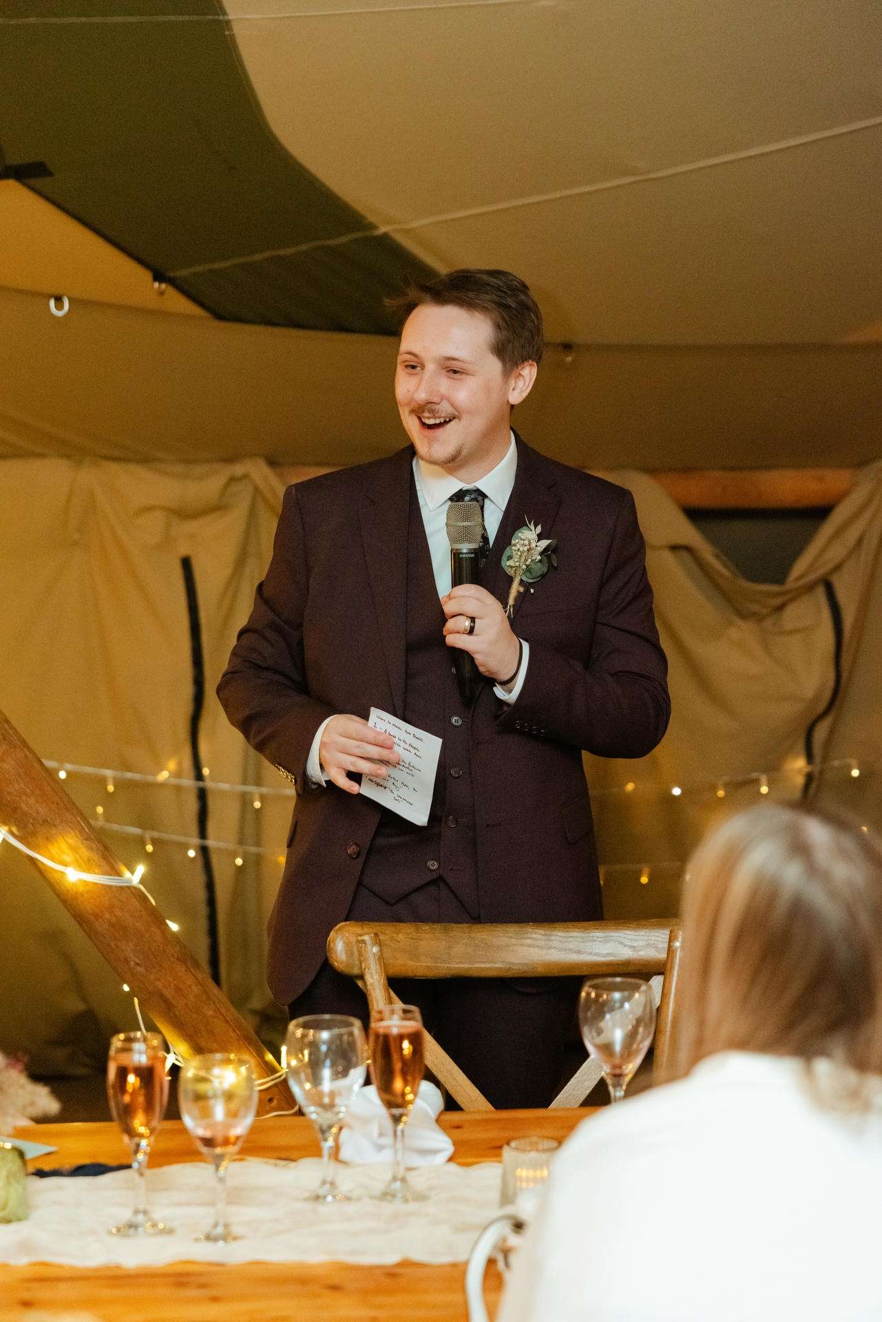 Man in a dark suit giving a speech at a wedding reception in a tent, holding a microphone and notes.