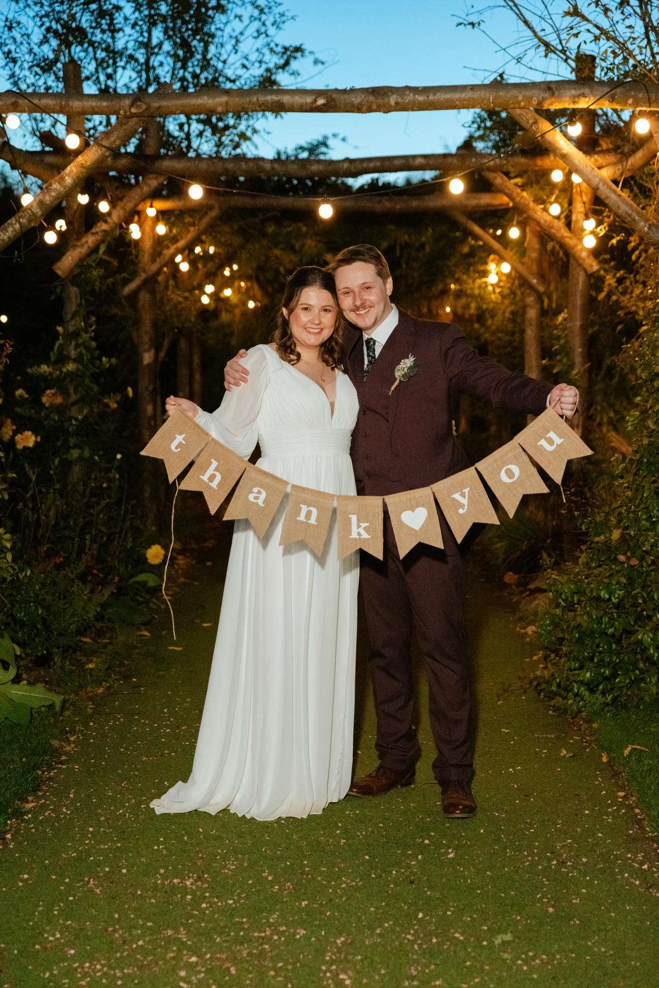 Bride and groom posing in a garden at dusk, holding a burlap banner that reads 'thank you'.