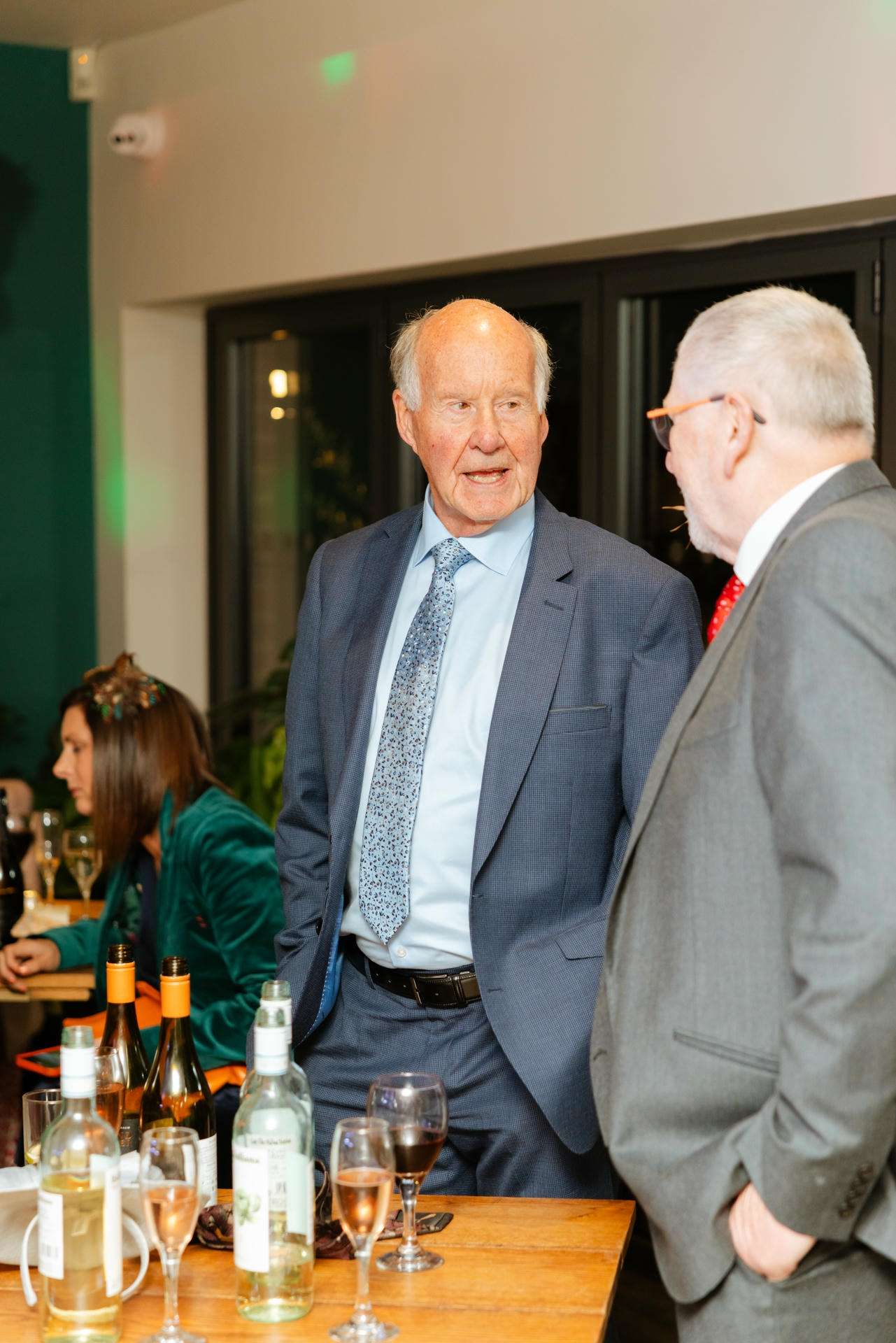 Two elderly men in suits stand and chat beside a wooden table cluttered with wine bottles and glasses at a social event.