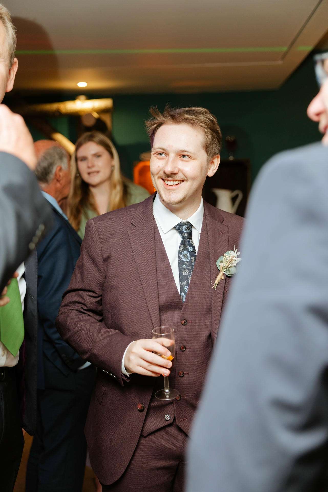 A man in a maroon suit smiles while holding a champagne flute at a wedding reception.