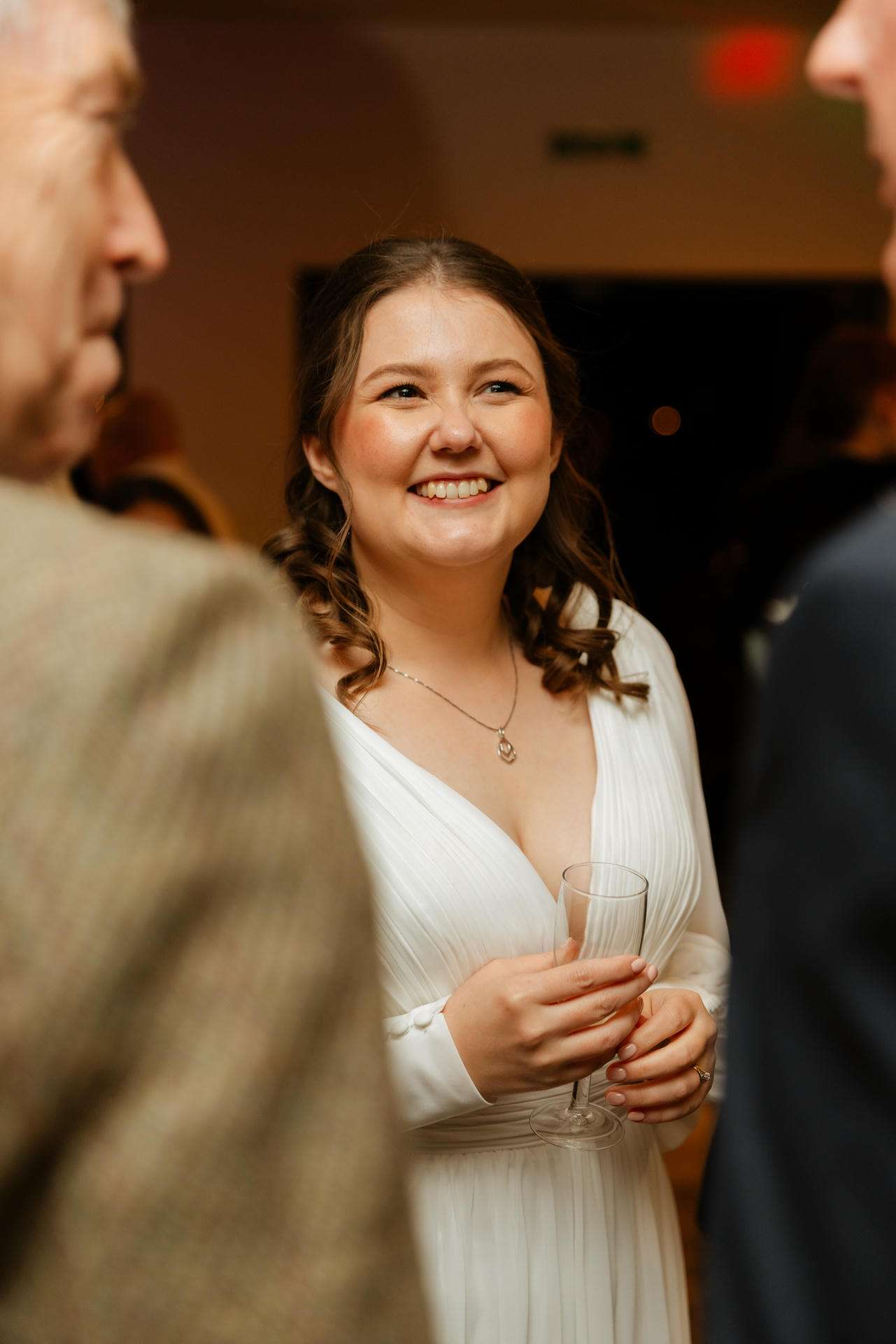 A smiling woman in a white dress holding a champagne flute at a social gathering.